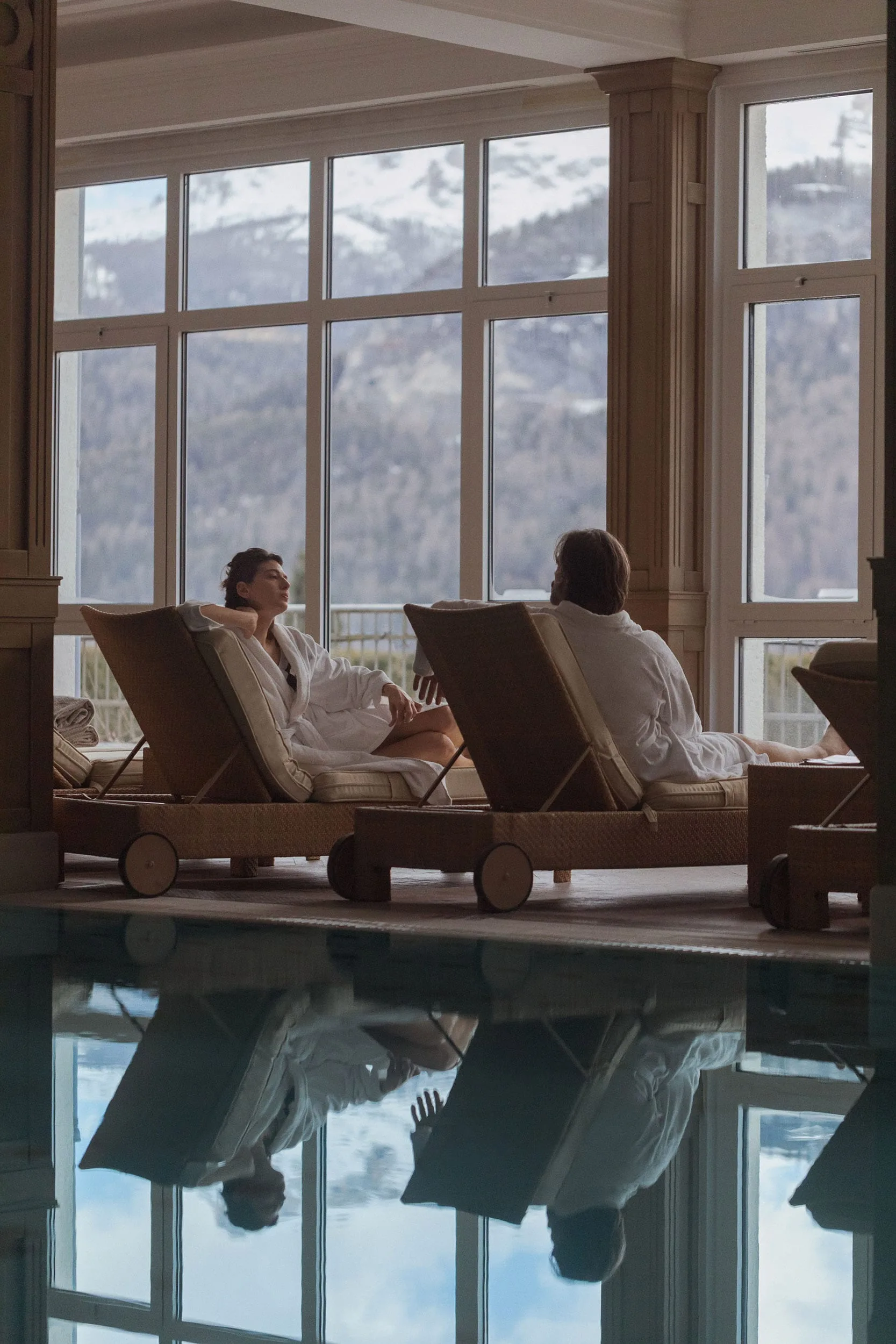Two guests in white robes recline on wicker loungers by an indoor pool with snowy mountain views, for Marriott.