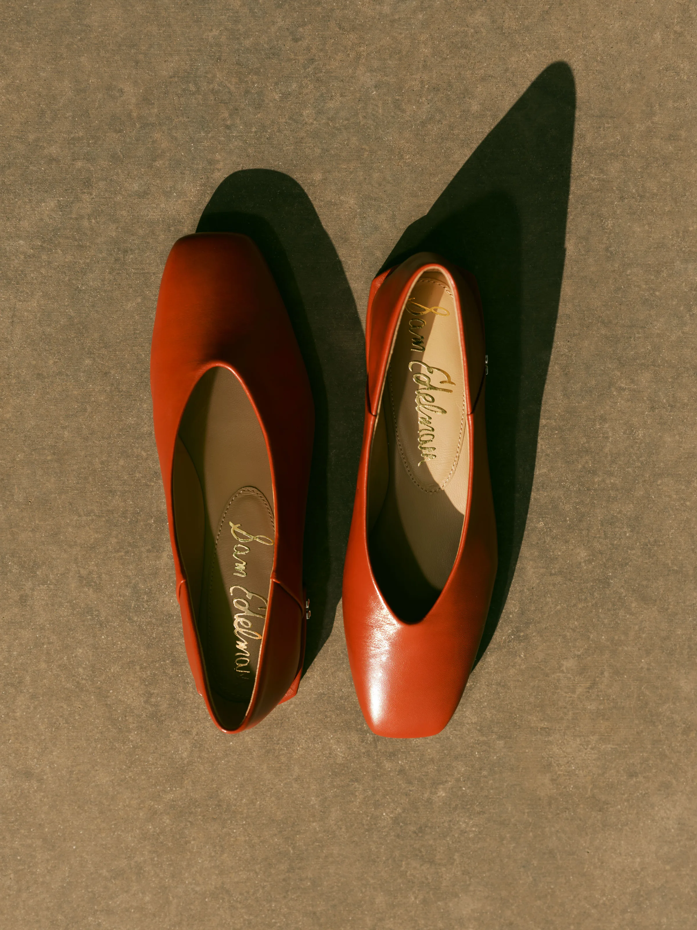 Pair of rust leather square-toe ballet flats casting hard shadows on a grey textured surface, for Sam Edelman.
