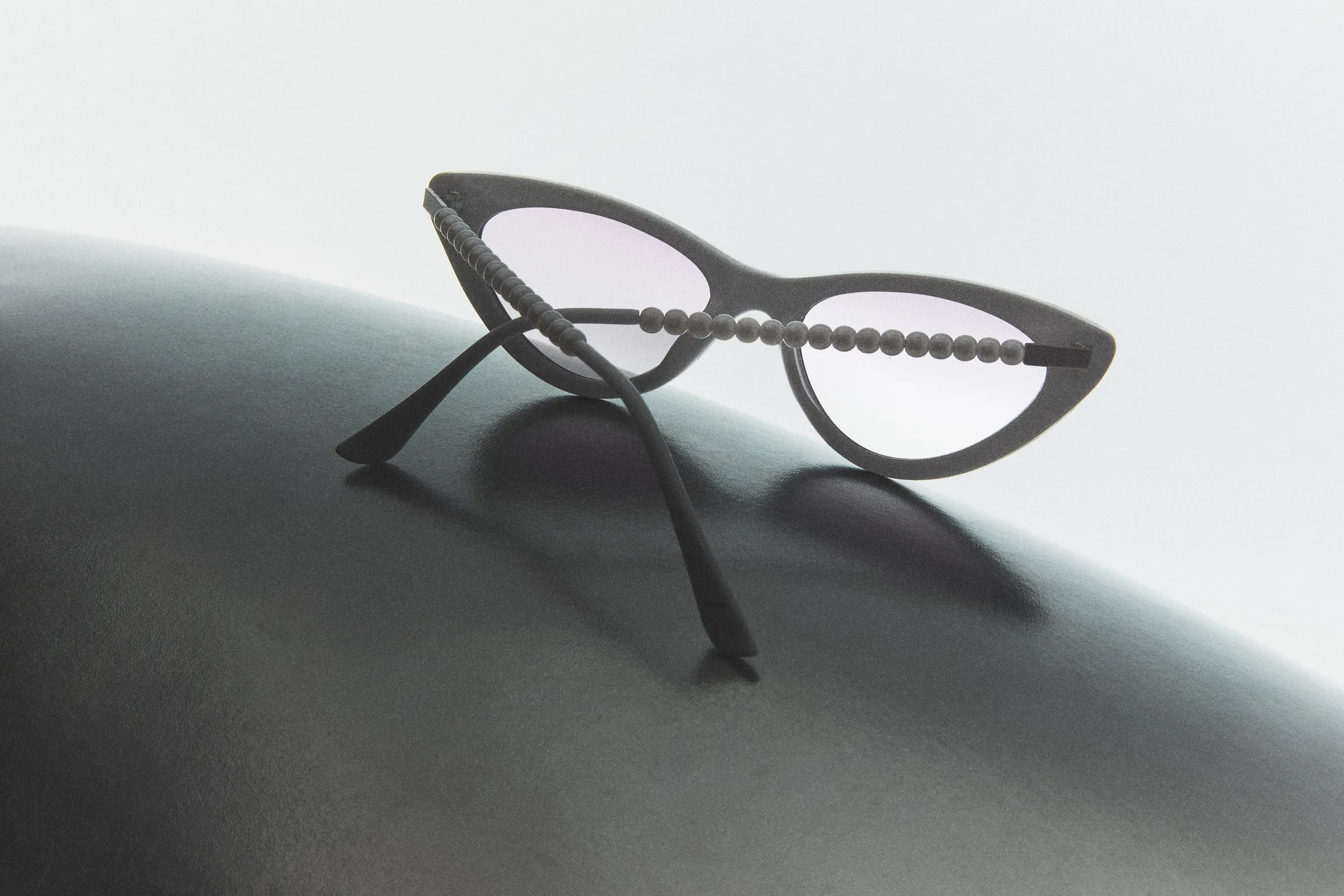 Grey cat-eye sunglasses with pearl-beaded temples resting on a dark curved surface against a pale backdrop, for Sam Edelman.