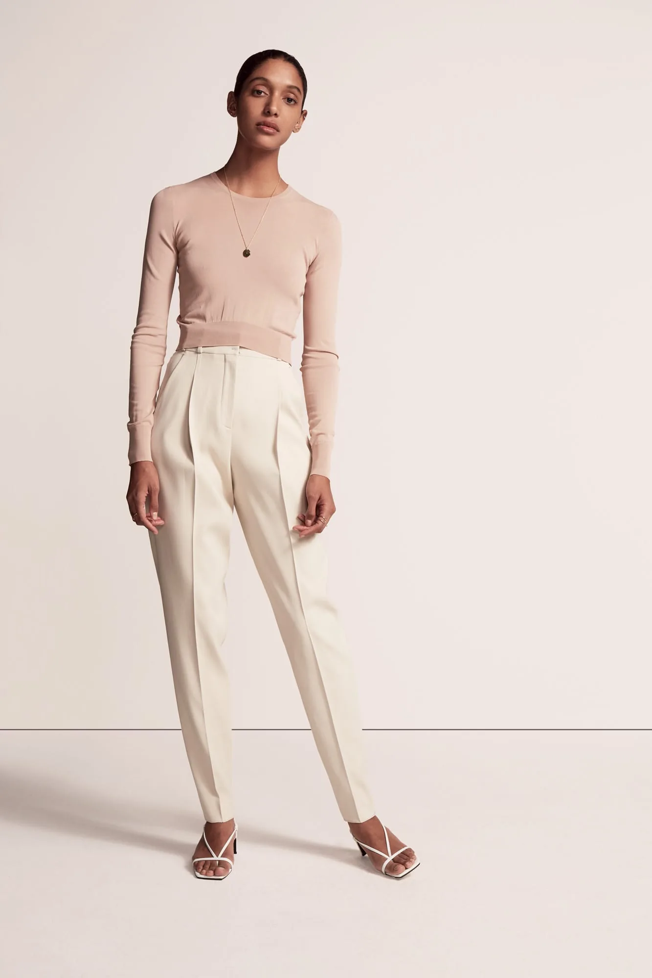 Model in a blush knit top, pleated ivory trousers and white strappy sandals on a pale backdrop, for Another Tomorrow.