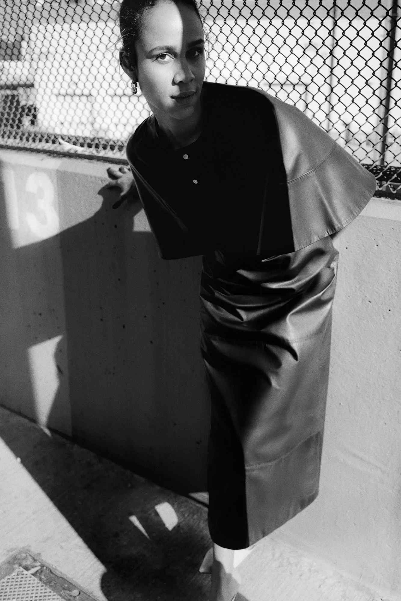 Black-and-white editorial of a model in a draped leather top and skirt by a chain-link fence, for The Last Magazine.
