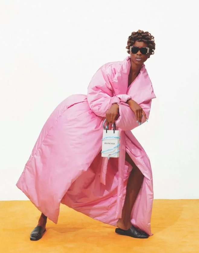 Model in an oversized pink puffer coat and sunglasses holding a small white Balenciaga bag on an orange floor, for Office Magazine.