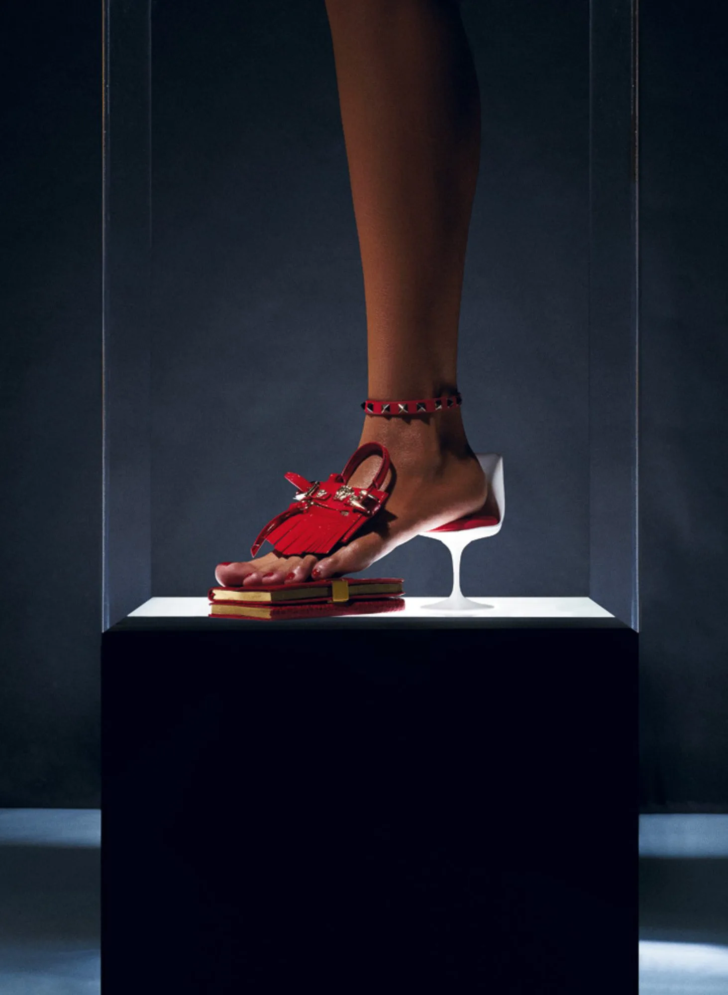 Bare foot wearing a red fringed studded sandal on red books beside a miniature white tulip chair in a lit vitrine, for Interview Magazine.