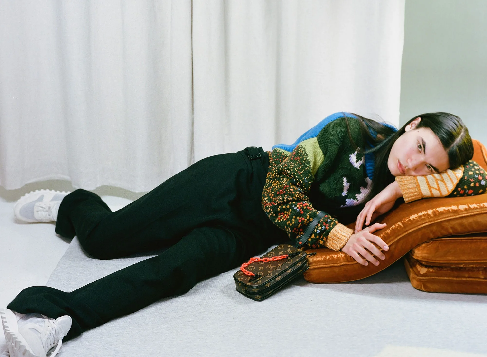 Model in a patterned knit sweater and black trousers lies on orange velvet cushions beside a monogram bag, for VMAN.