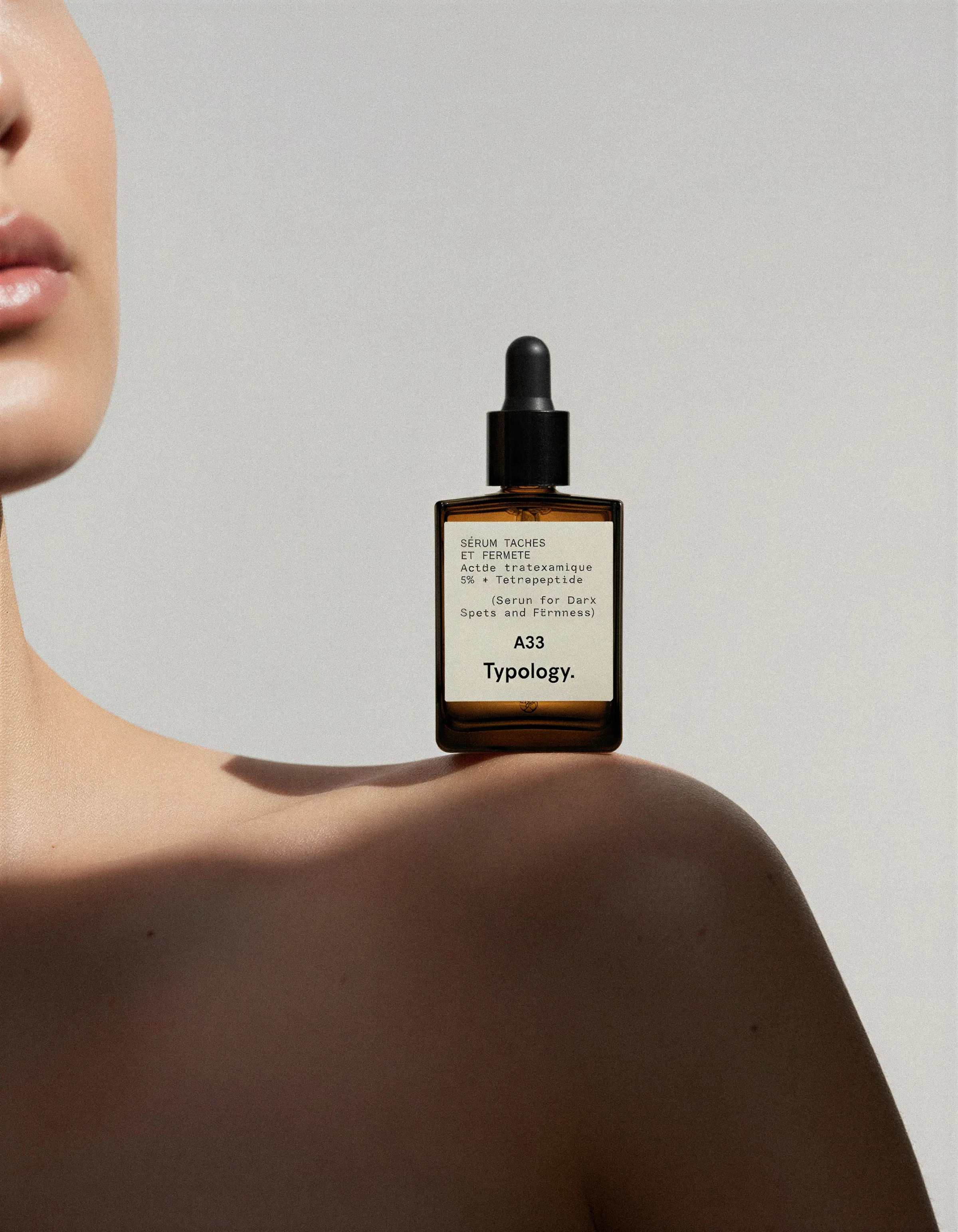 AI image of an amber Typology dropper serum bottle balanced on a model's bare shoulder against a grey backdrop. Typology AI Concept.