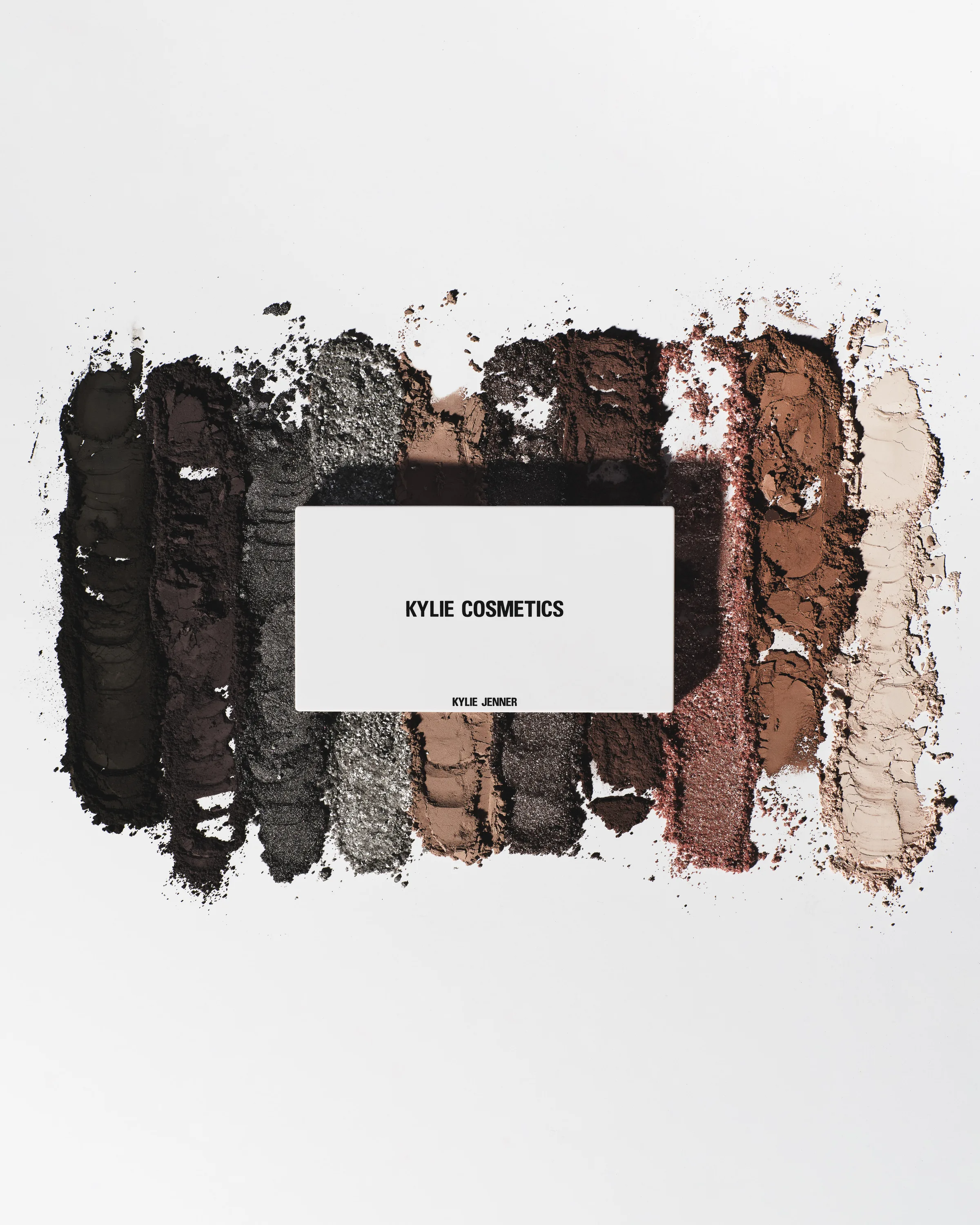 White eyeshadow palette over crushed swatches of black, grey, brown and nude shadows on a white background, for Kylie Cosmetics.