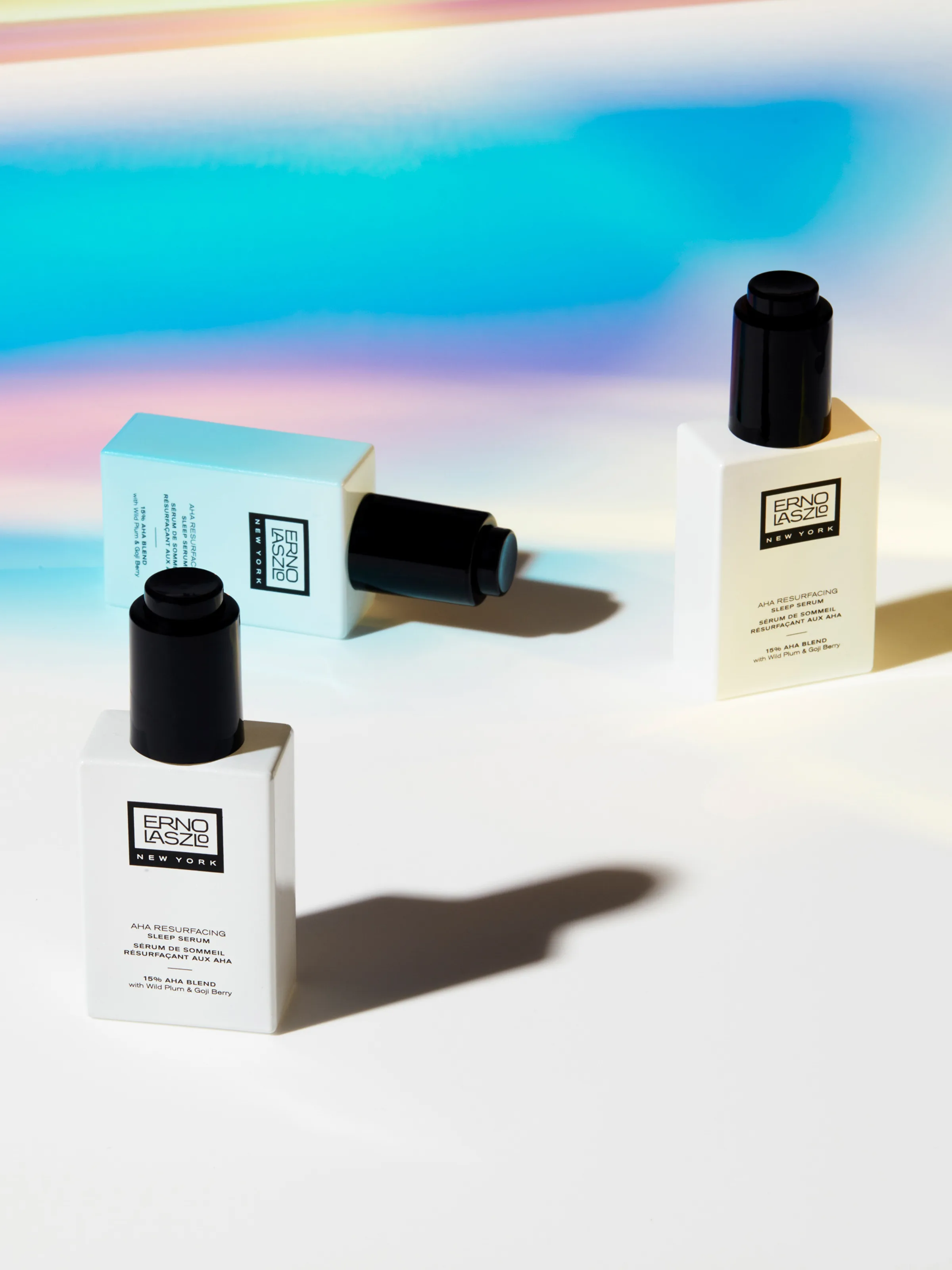 Three AHA Resurfacing Sleep Serum bottles with black droppers on white under iridescent rainbow light, for Erno Laszlo.