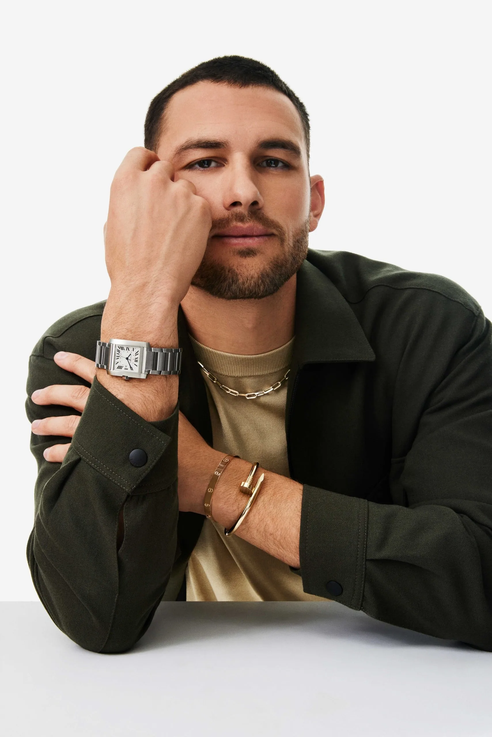 Man in olive jacket resting head on fist, wearing a steel Tank watch, chain necklace and gold Juste un Clou bracelet, for Cartier.