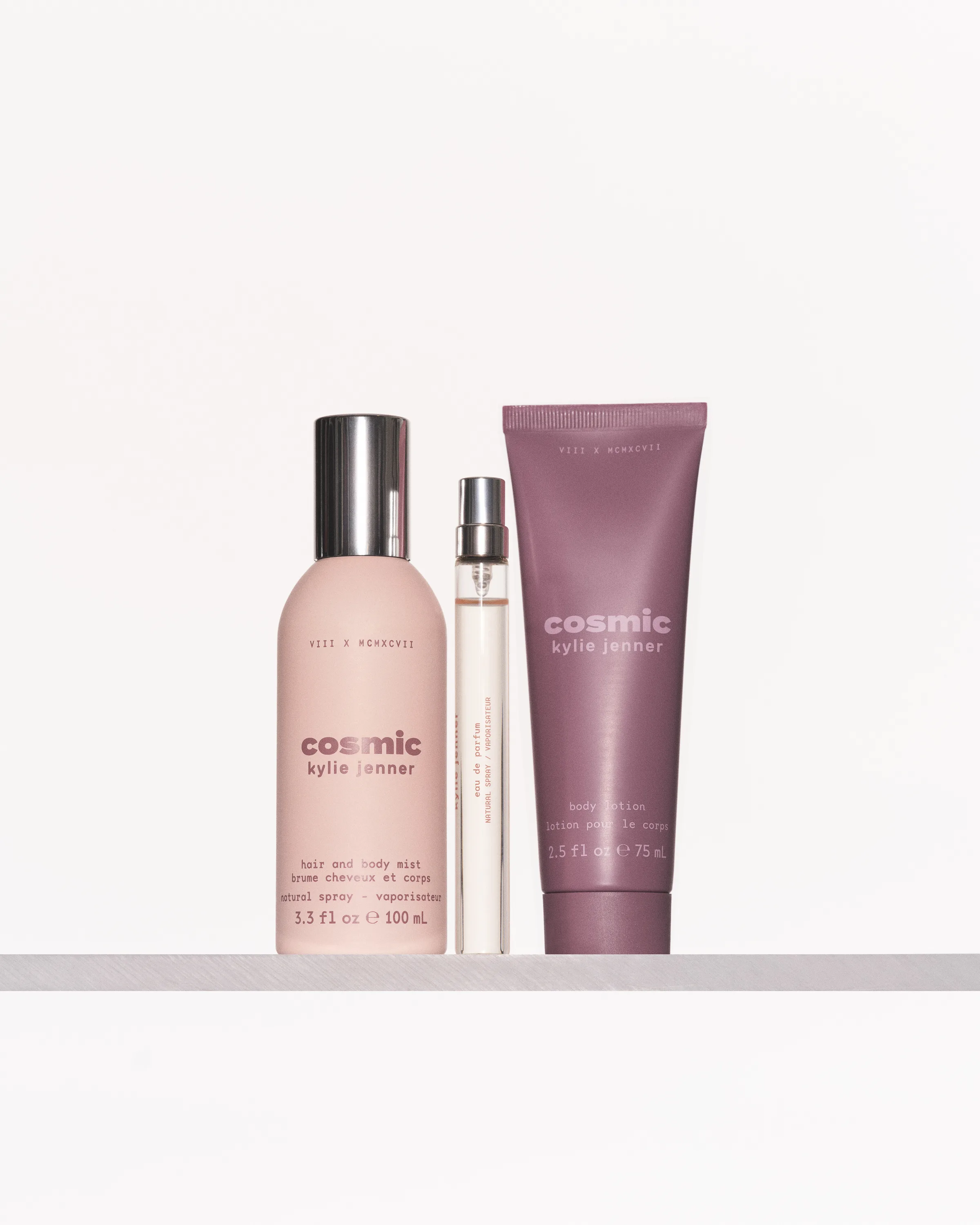 Pink Cosmic hair and body mist, eau de parfum travel spray and mauve body lotion on a white shelf, for Kylie Jenner.