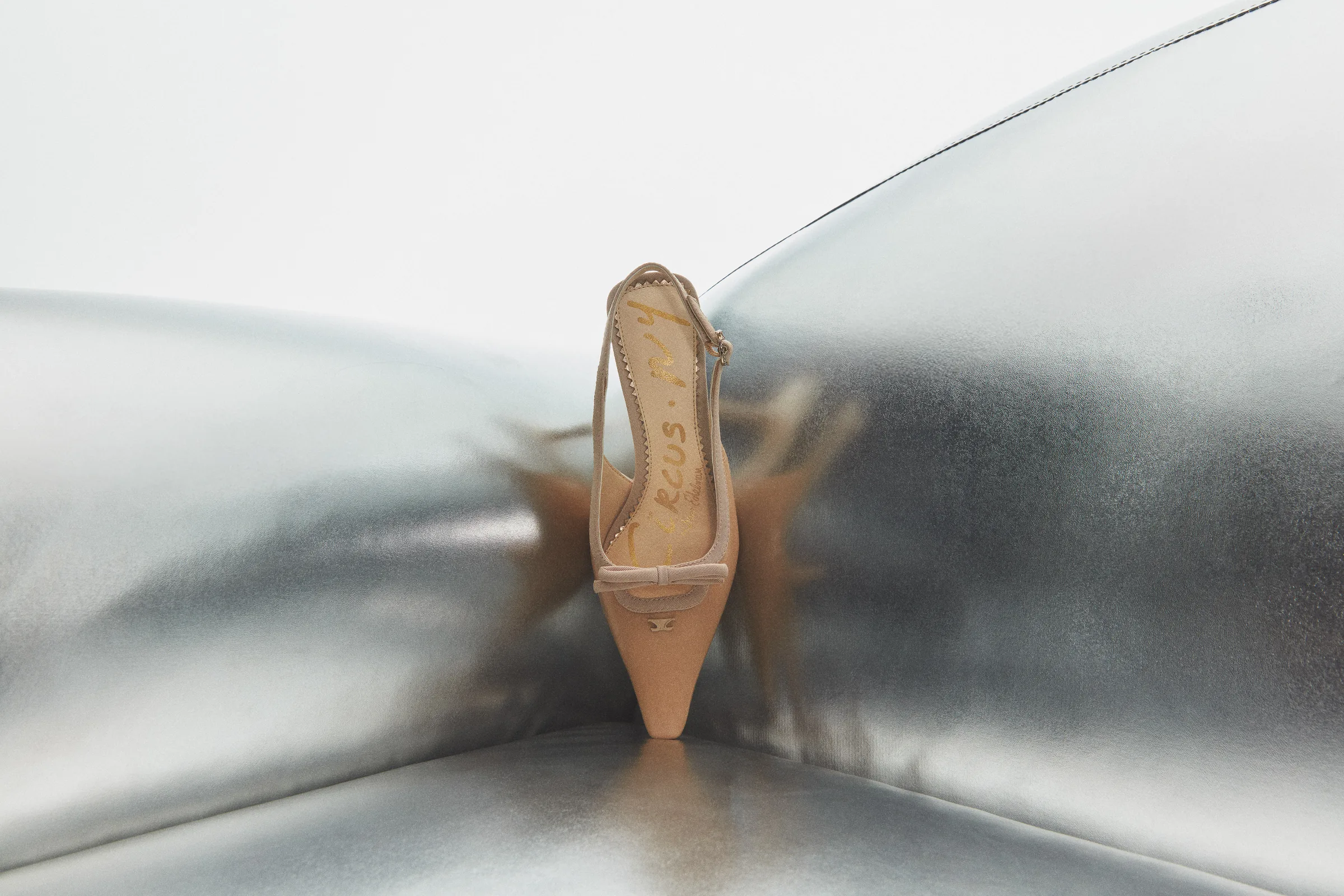 Nude pointed-toe slingback heel with a bow, perched on a silver metallic cushioned surface, for Sam Edelman.