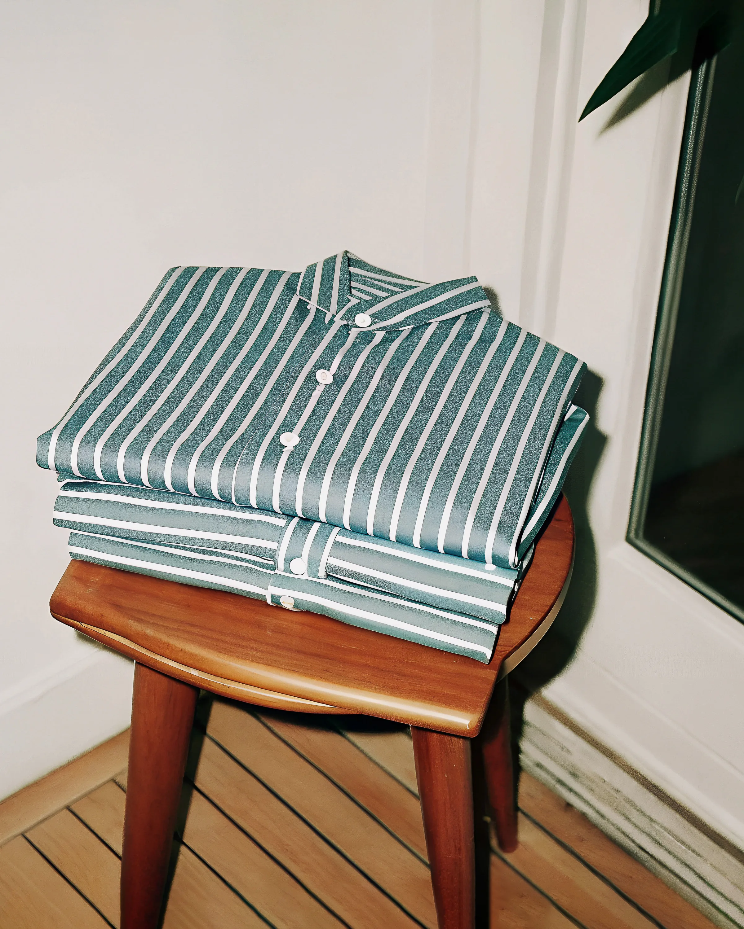Stack of folded teal and white striped shirts on a wooden stool beside a window. Massimo Dutti AI Concept.