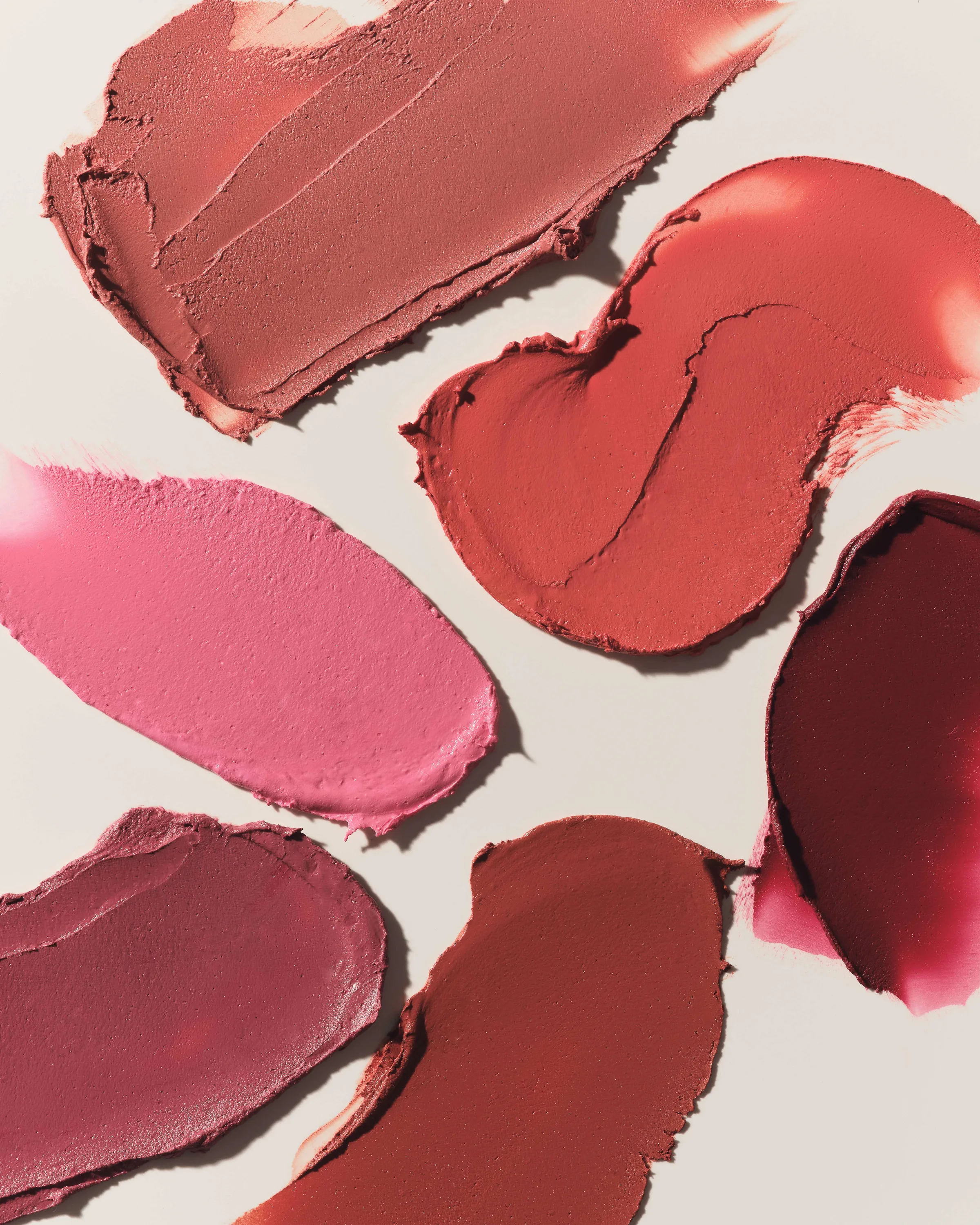 Six lipstick swatches in pink, rose, red and plum smeared across a cream background in sunlight, for Kylie Cosmetics.