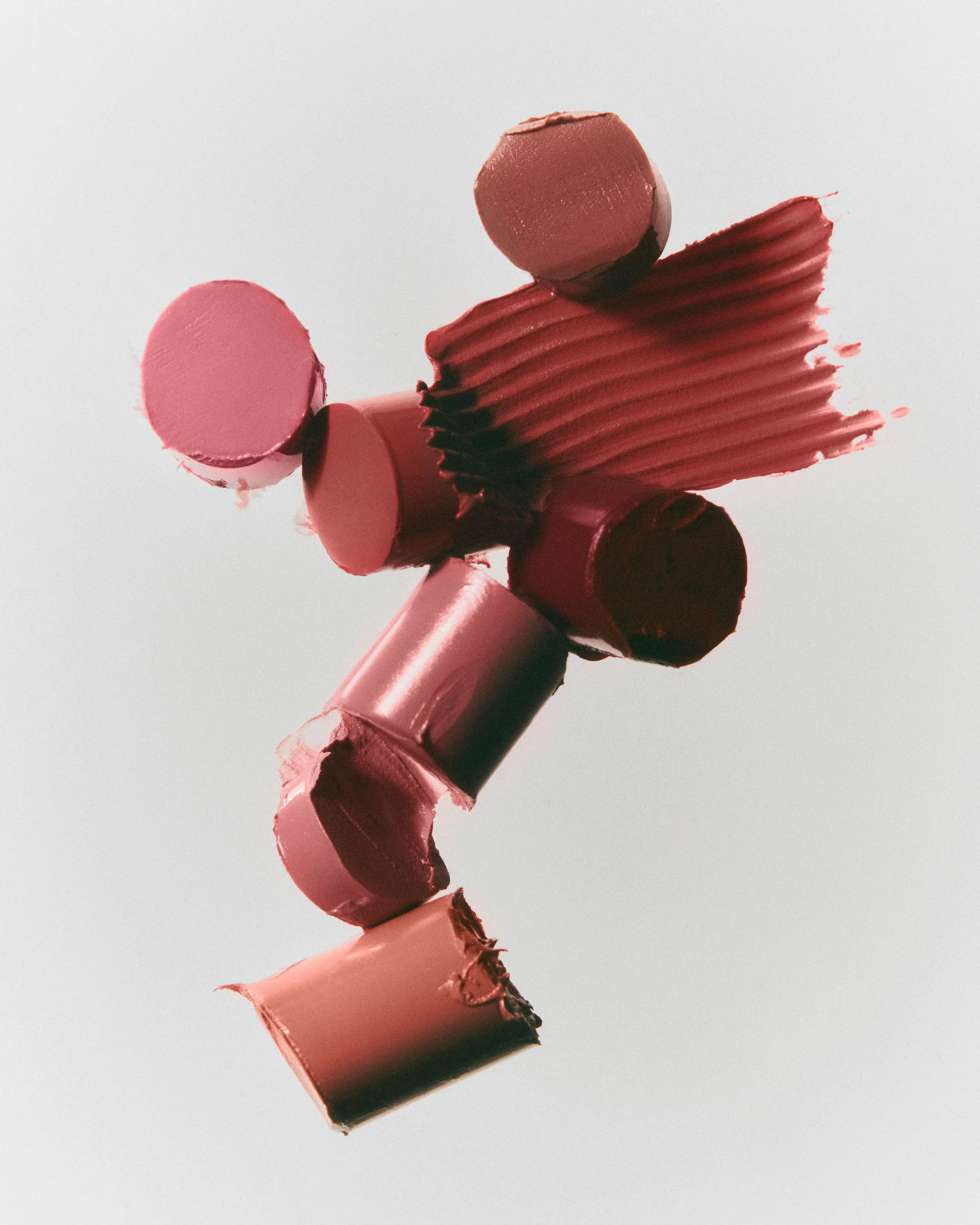 Still life of cut lipstick bullets in pink and nude shades with a textured red swatch on grey, for Kylie Cosmetics.