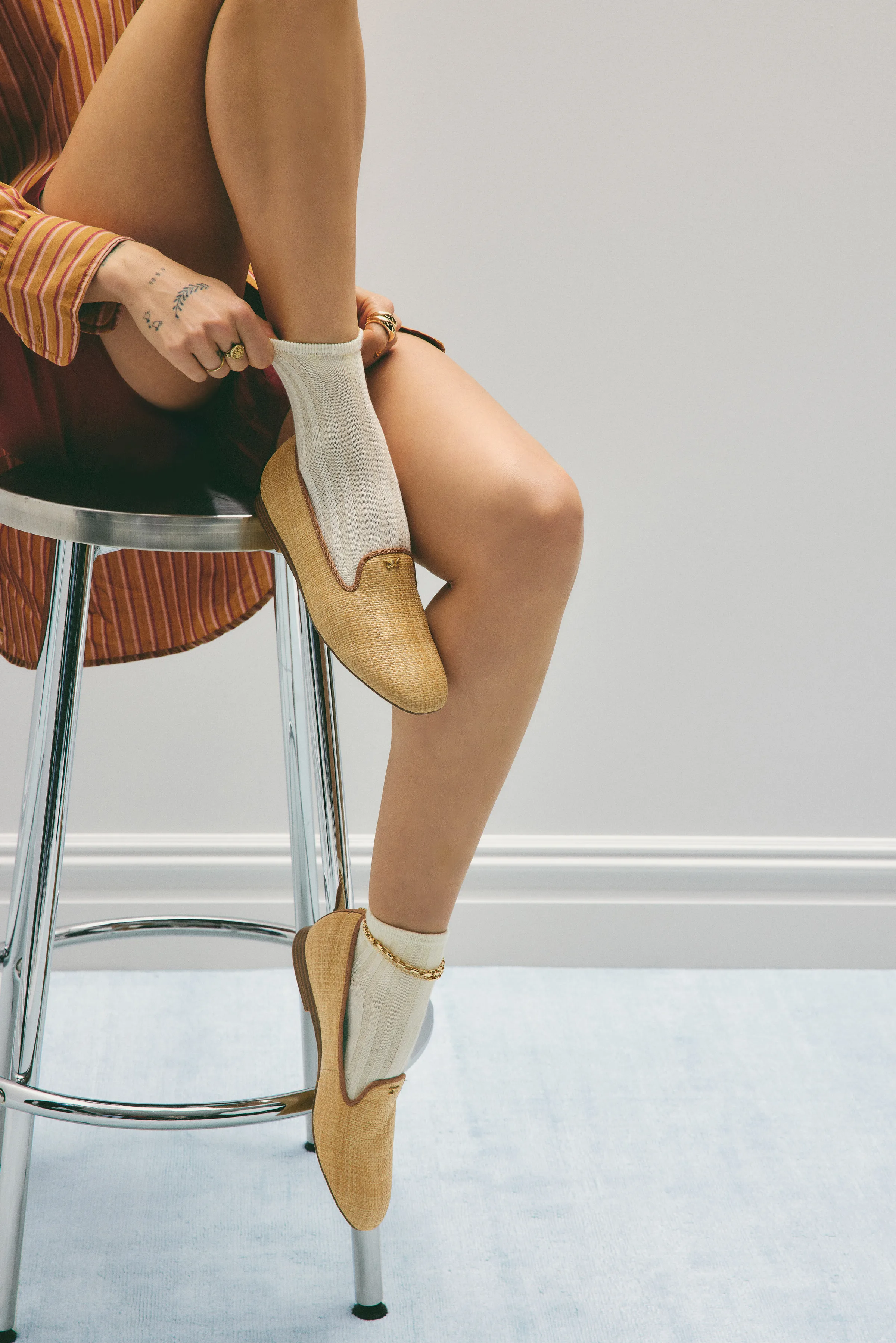 Model on a chrome stool wearing natural raffia loafers, cream ankle socks and a gold anklet, for Sam Edelman.