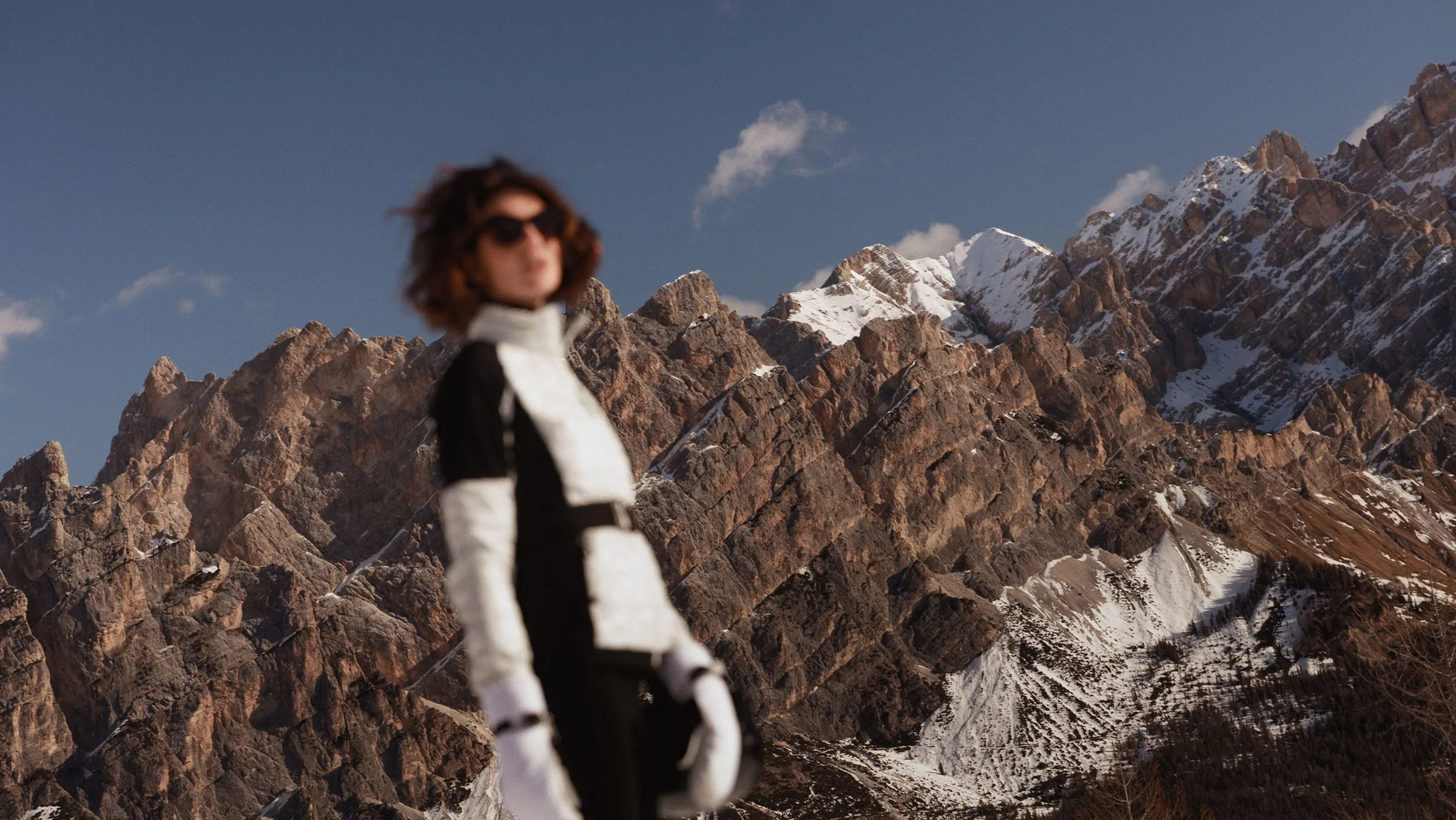 Woman in sunglasses and black and white ski suit, blurred before snowy rocky mountain peaks, for Marriott.