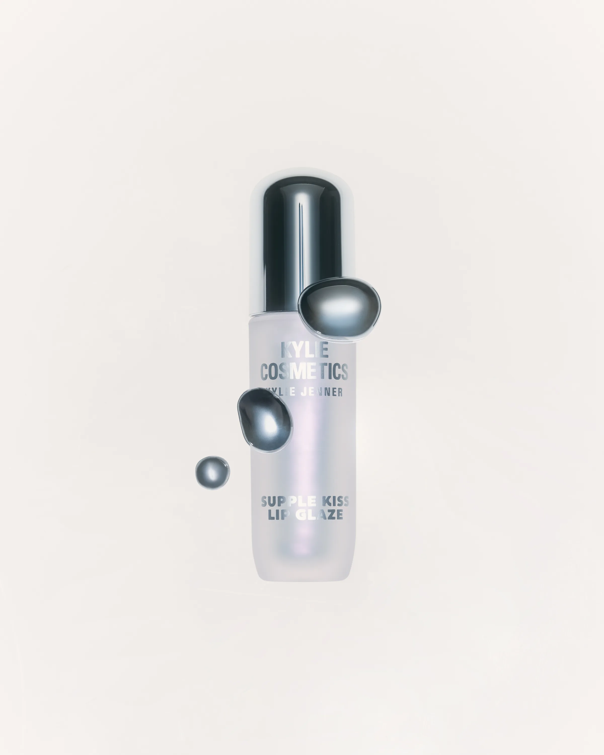 Iridescent Supple Kiss Lip Glaze tube with chrome cap and floating silver droplets on a white background, for Kylie Cosmetics.