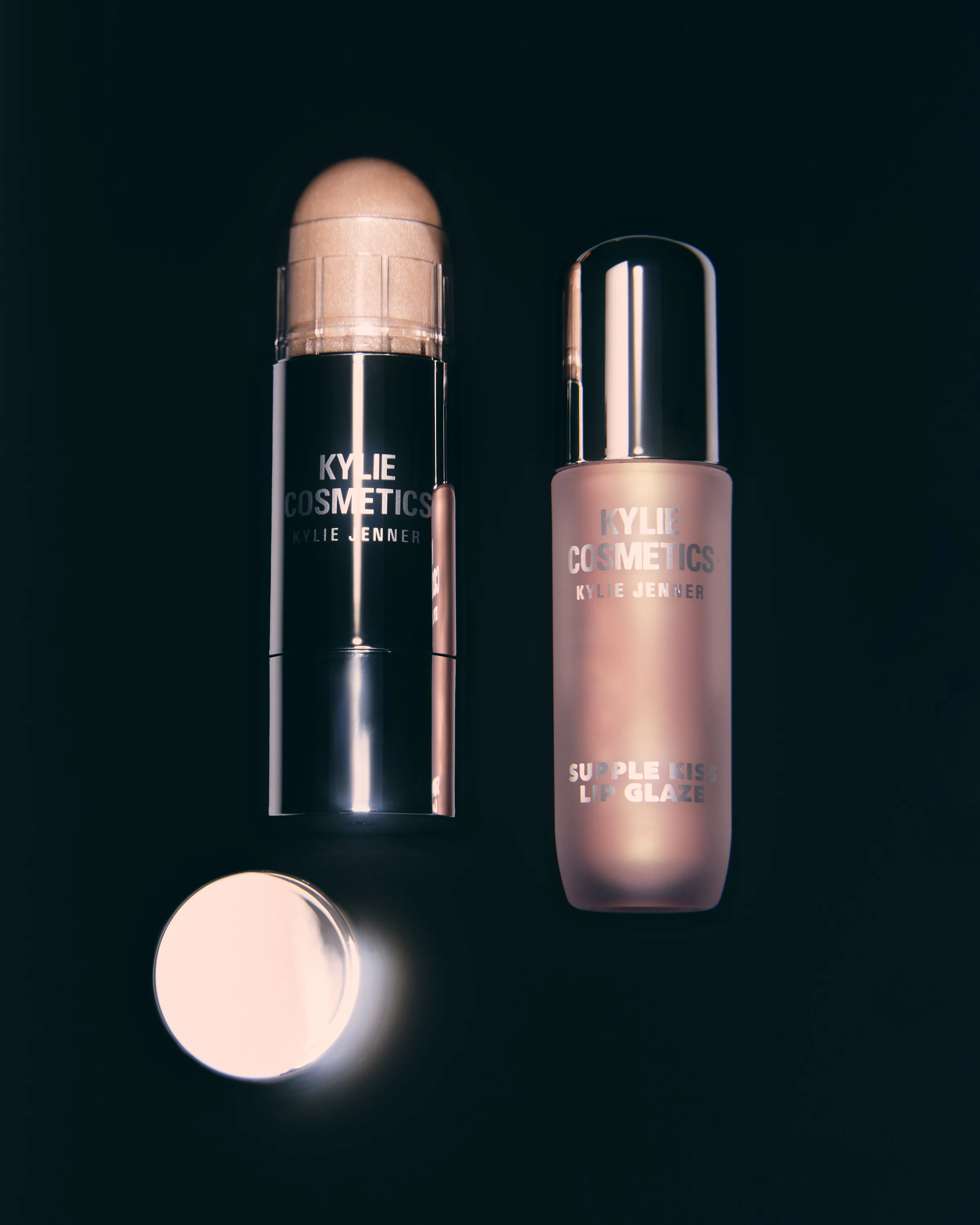 Still life of a shimmering balm stick, its cap and a pink lip glaze bottle on a black background, for Kylie Cosmetics.