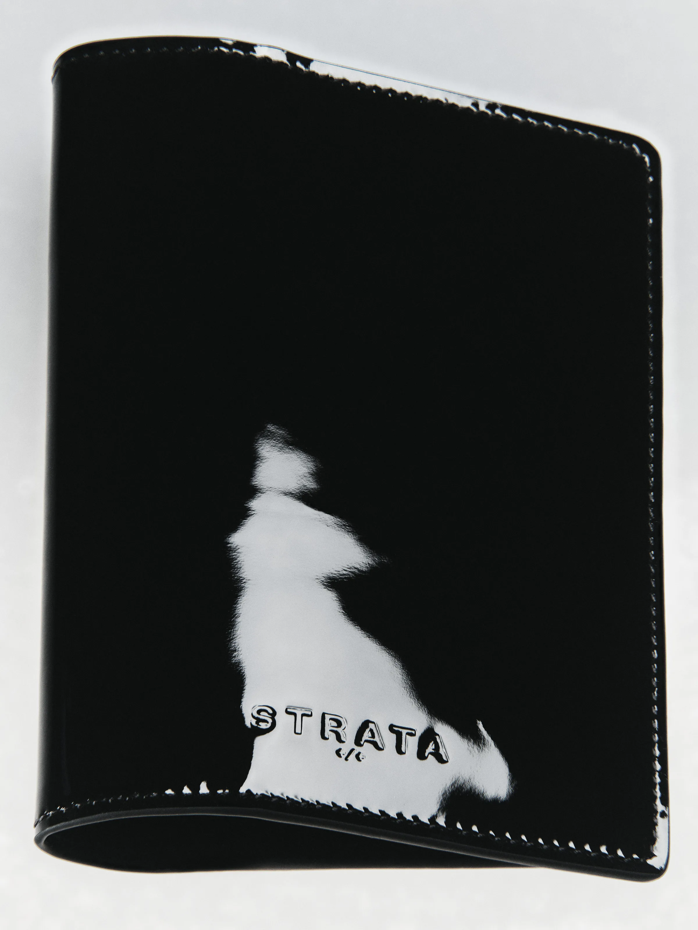 Black patent leather travel sleeve with embossed logo and glossy reflection on a pale grey backdrop, for Strata.