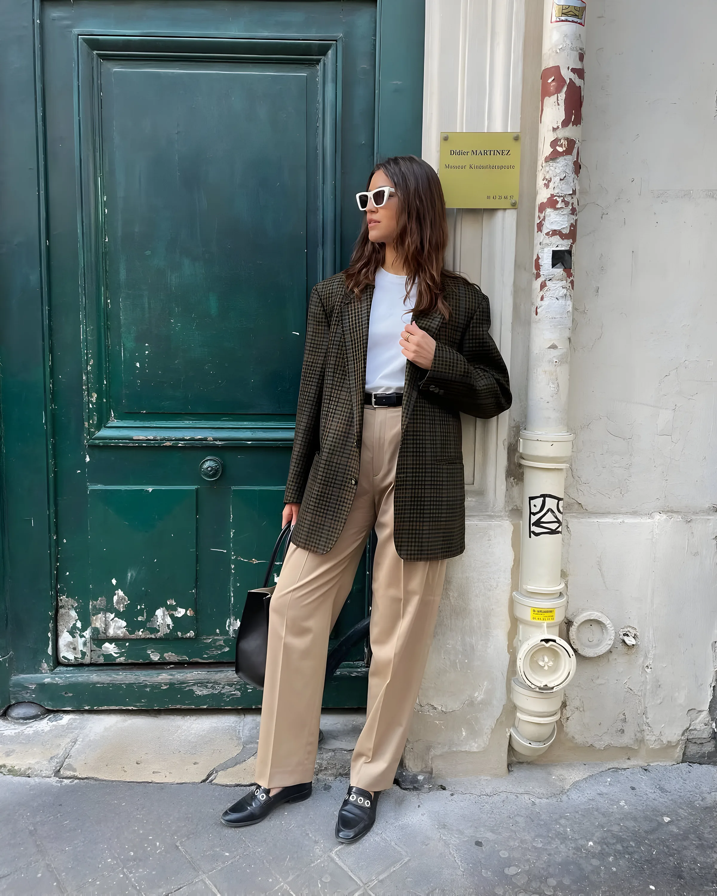 Woman in a check blazer, white tee and beige trousers leaning against a green door on a city street, wearing white sunglasses.