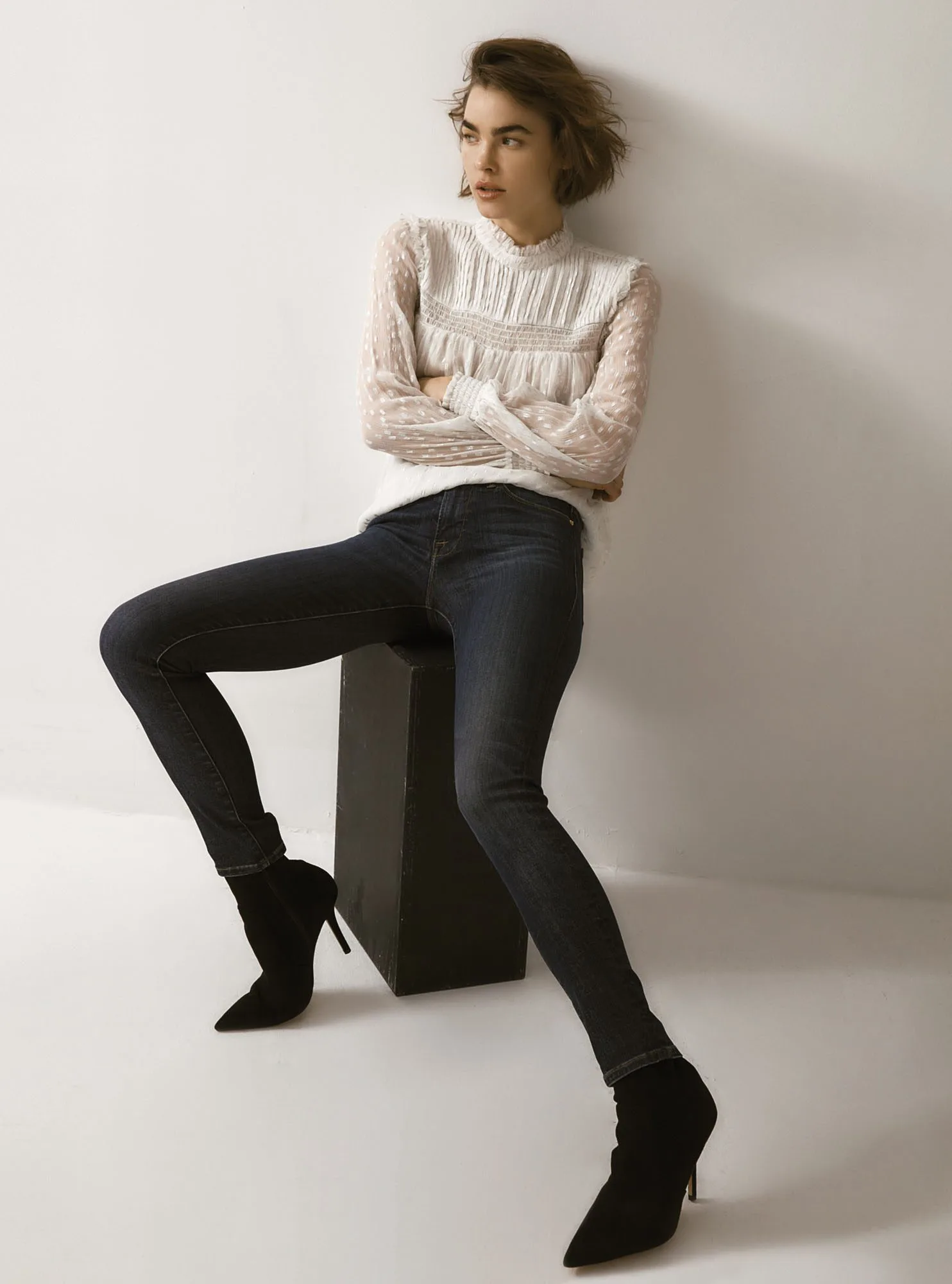 Model in a sheer white lace blouse and dark skinny jeans sits on a black box in suede ankle boots, for FRAME.