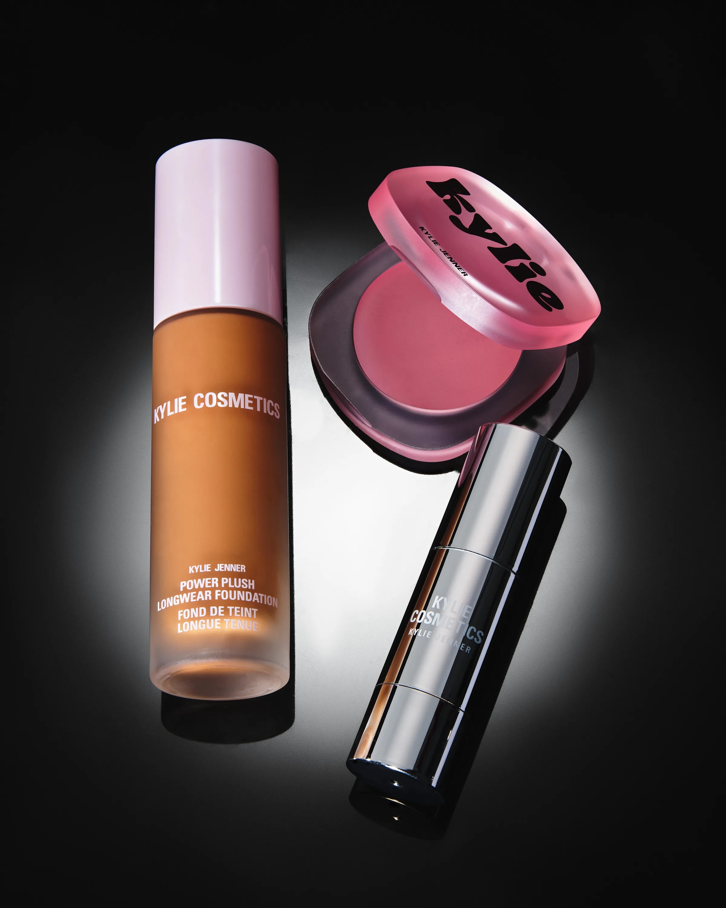 Still life of foundation bottle, pink glow balm compact and silver dewy stick under a spotlight on black, for Kylie Cosmetics.