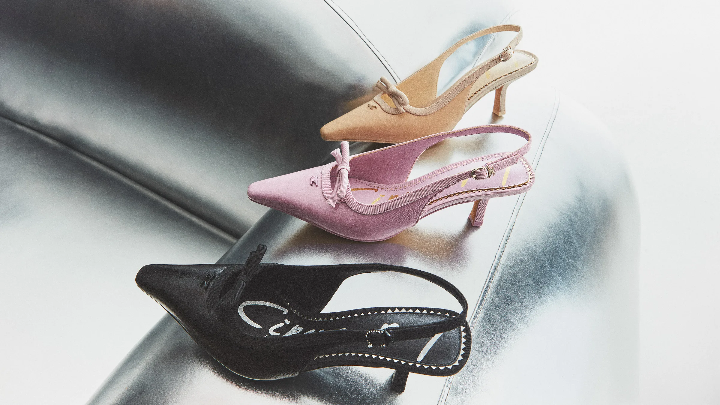 Black, pink and vanilla Cassidy slingback heels with bows arranged on a curved chrome surface, for Circus NY.