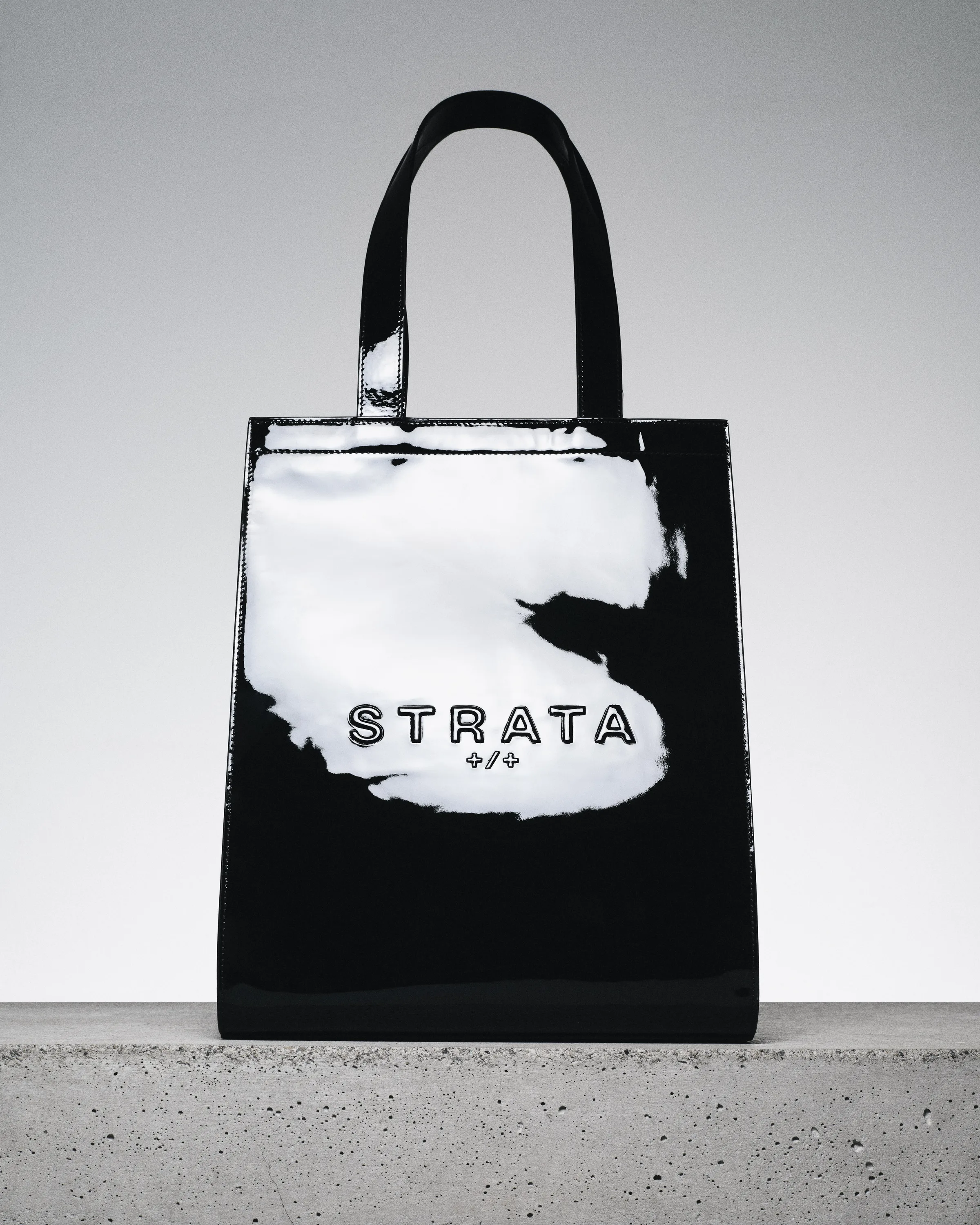 Black patent leather Carmen tote with Strata logo on a concrete ledge against a grey backdrop, for Strata.