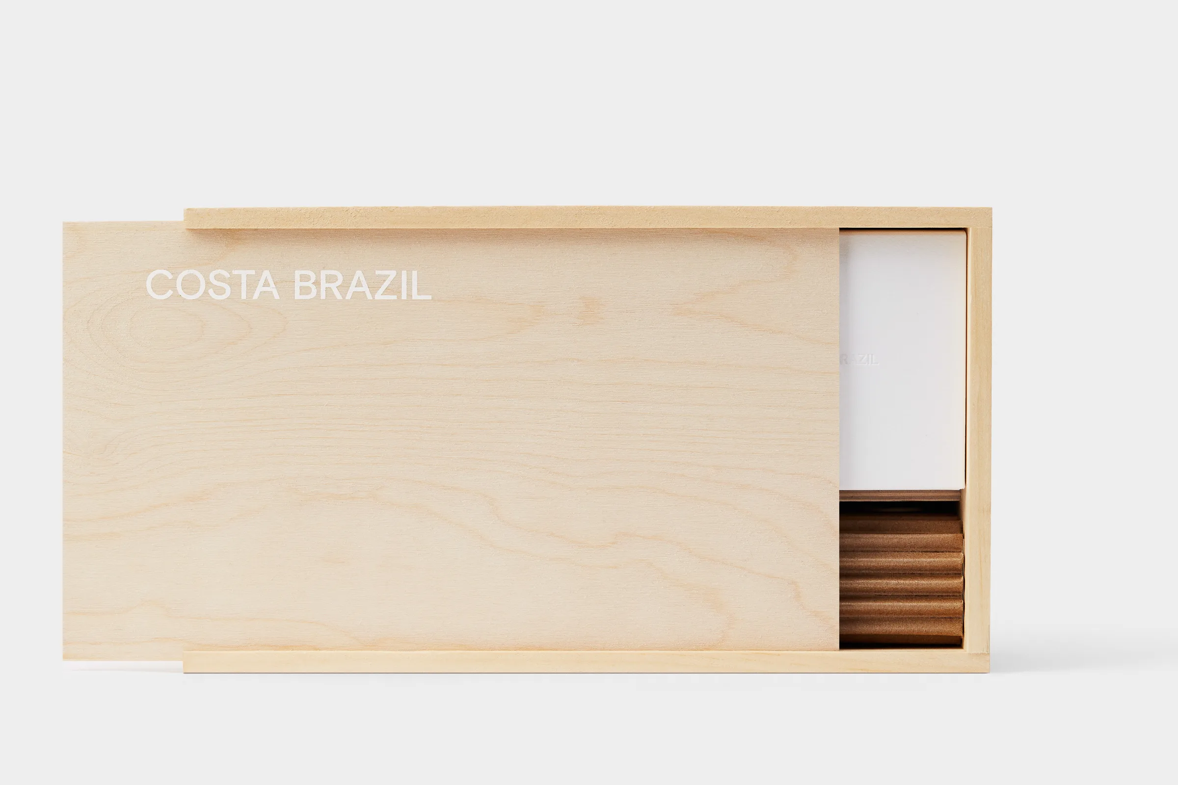 Light maple wooden box with sliding lid partly open, showing white card and wooden pieces, on grey, for Costa Brazil.