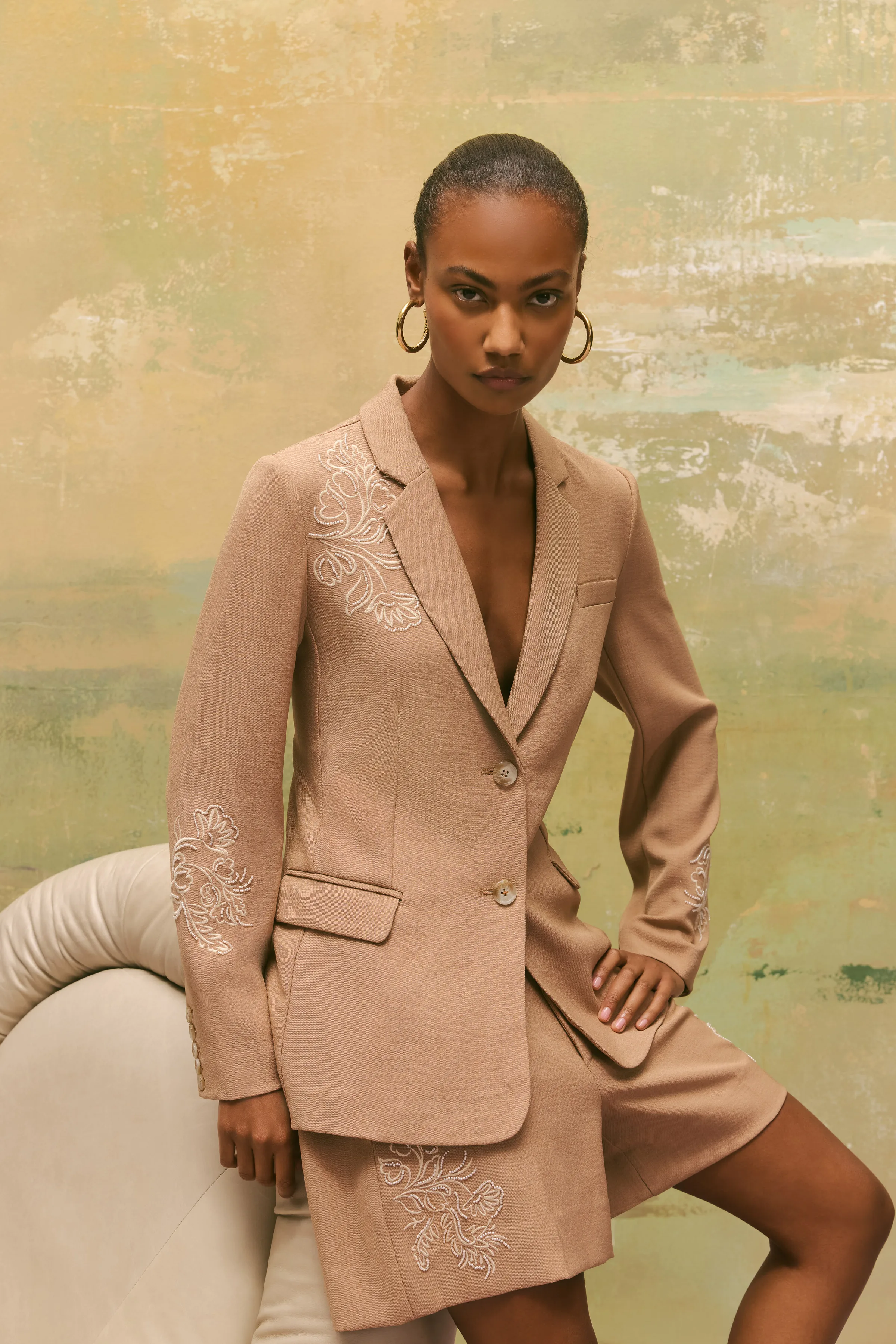 Model in a tan blazer and shorts with white floral embroidery, posed against a painted backdrop, for Sam Edelman.