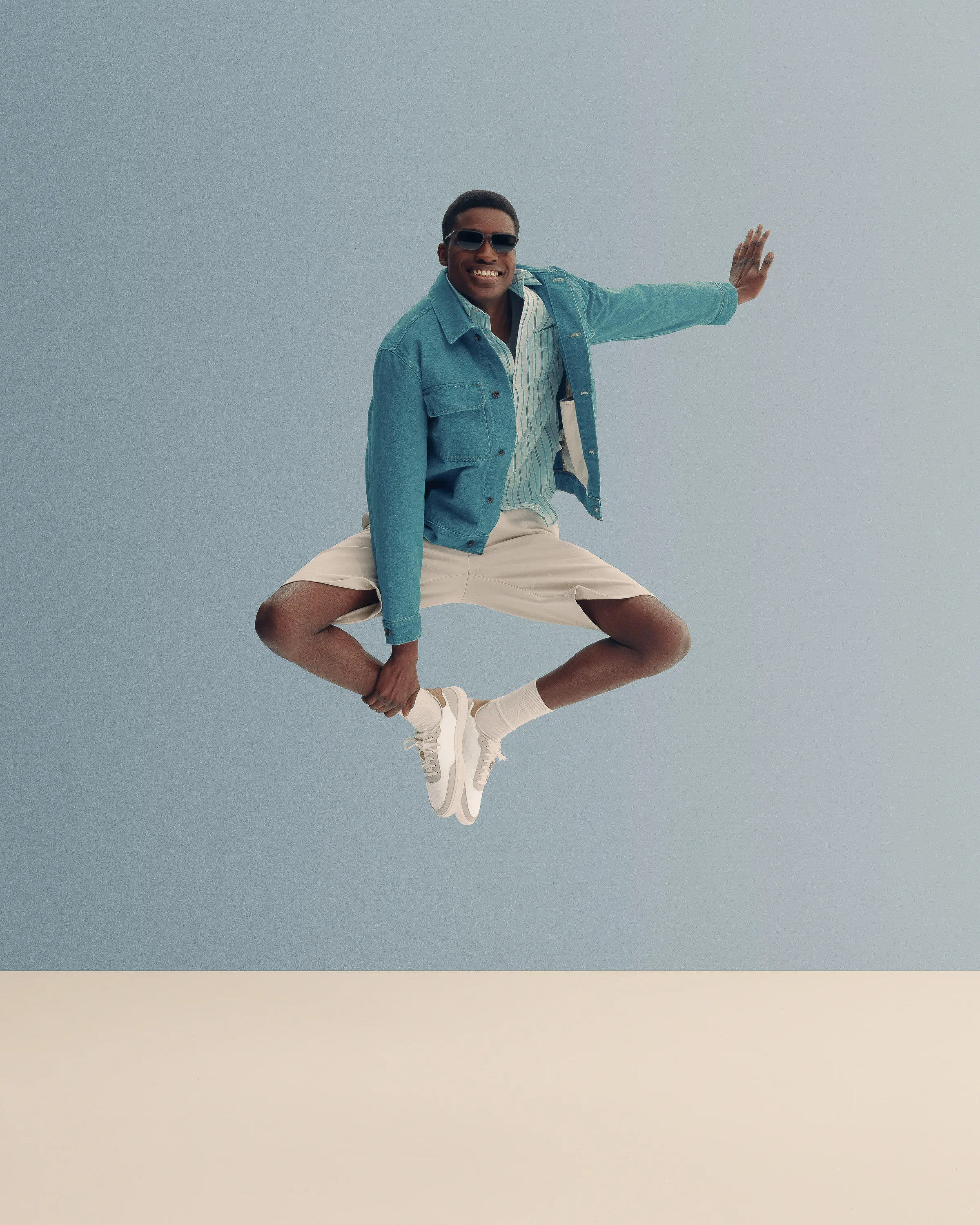 Smiling man mid-jump in teal denim jacket, cream shorts and white sneakers against a pale blue backdrop, for Cole Haan.
