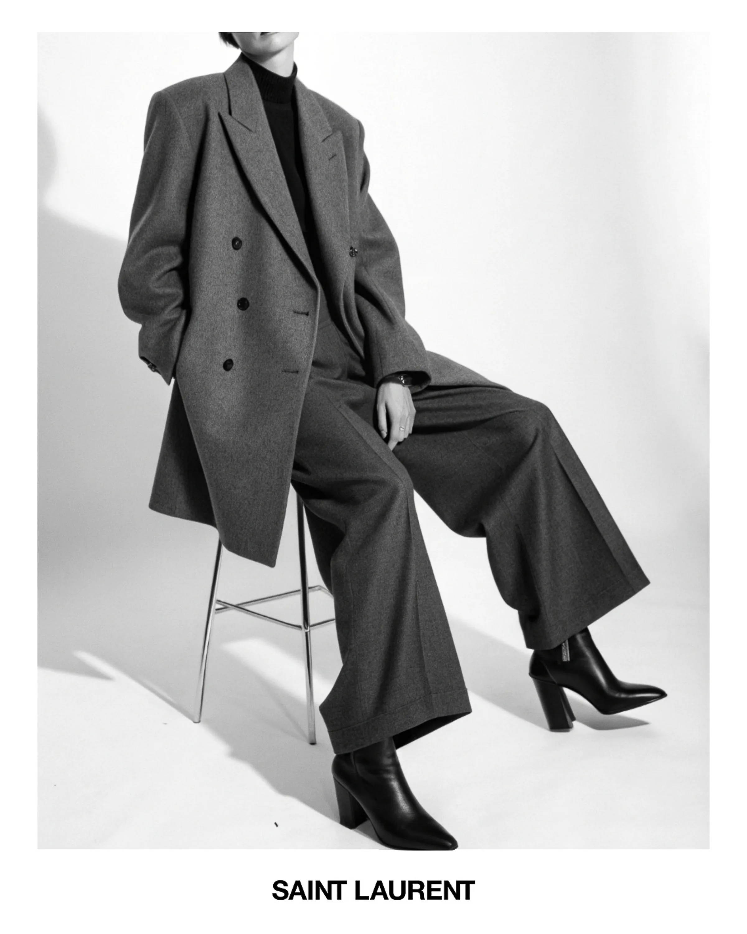 AI image of a model in a grey double-breasted coat, wide trousers and black heeled boots seated on a stool. Saint Laurent AI Concept.