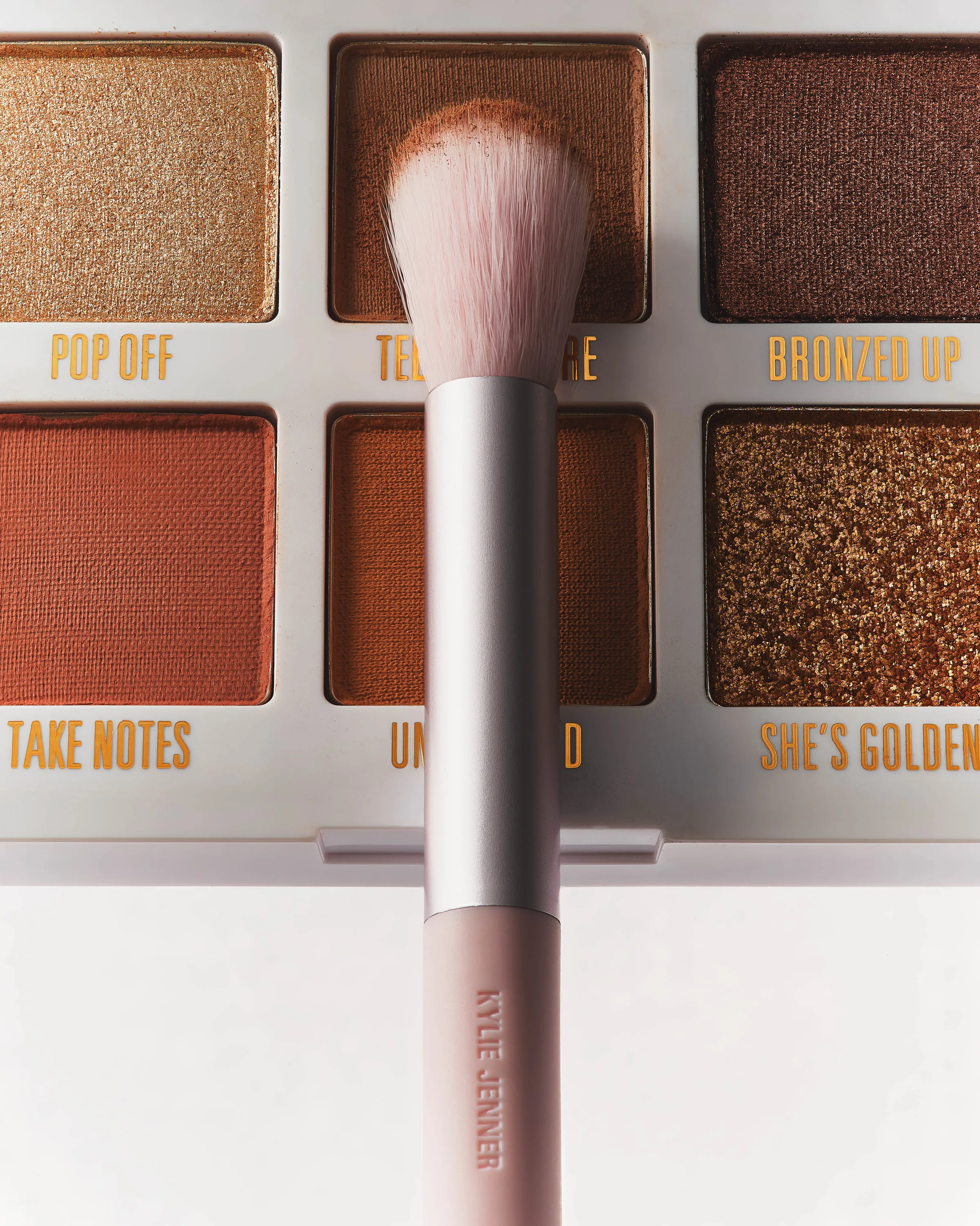 Pink makeup brush held over a bronze and gold eyeshadow palette with named shades, for Kylie Cosmetics.