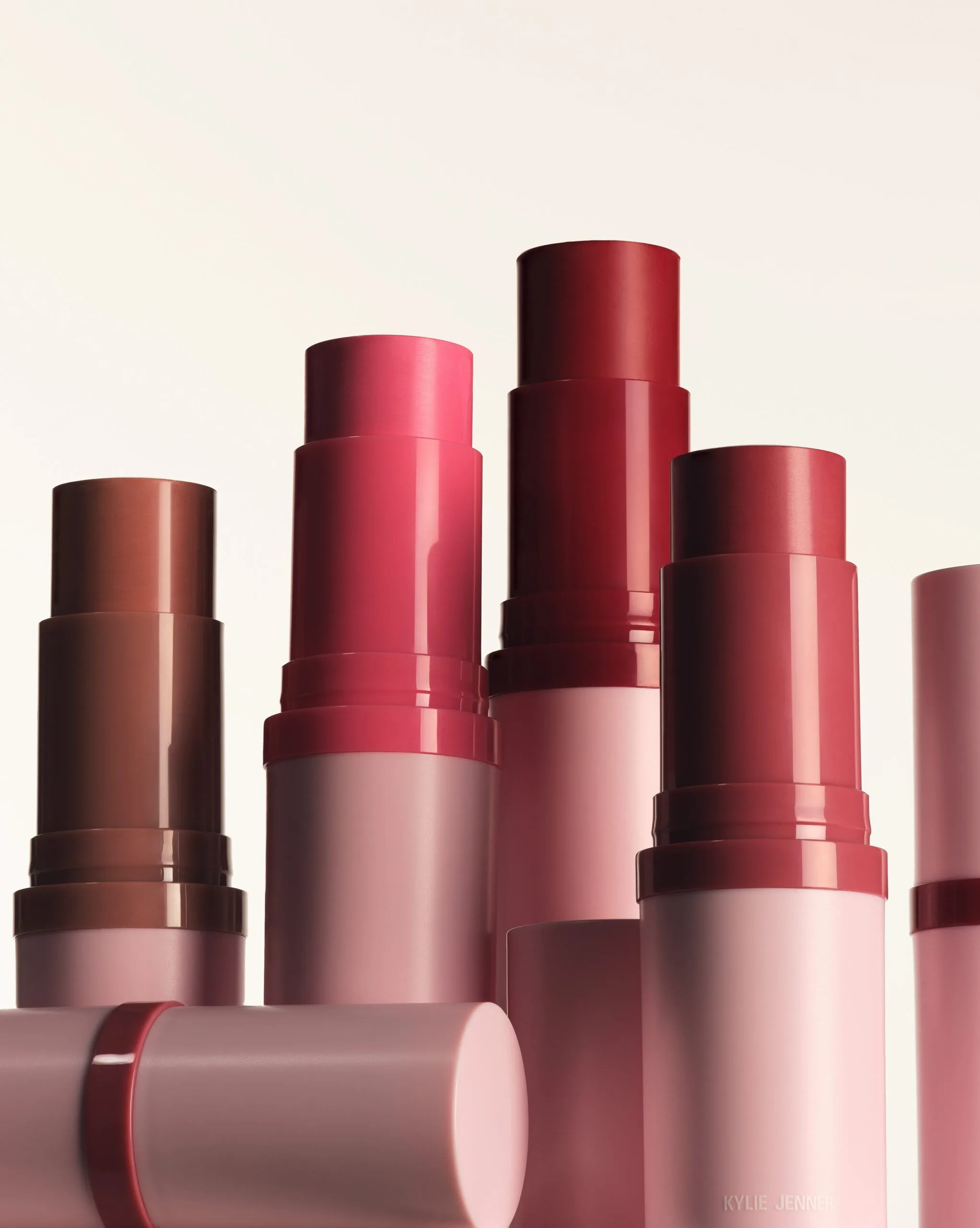 Uncapped blush sticks in brown, pink and berry shades with pink tubes on a cream background, for Kylie Cosmetics.