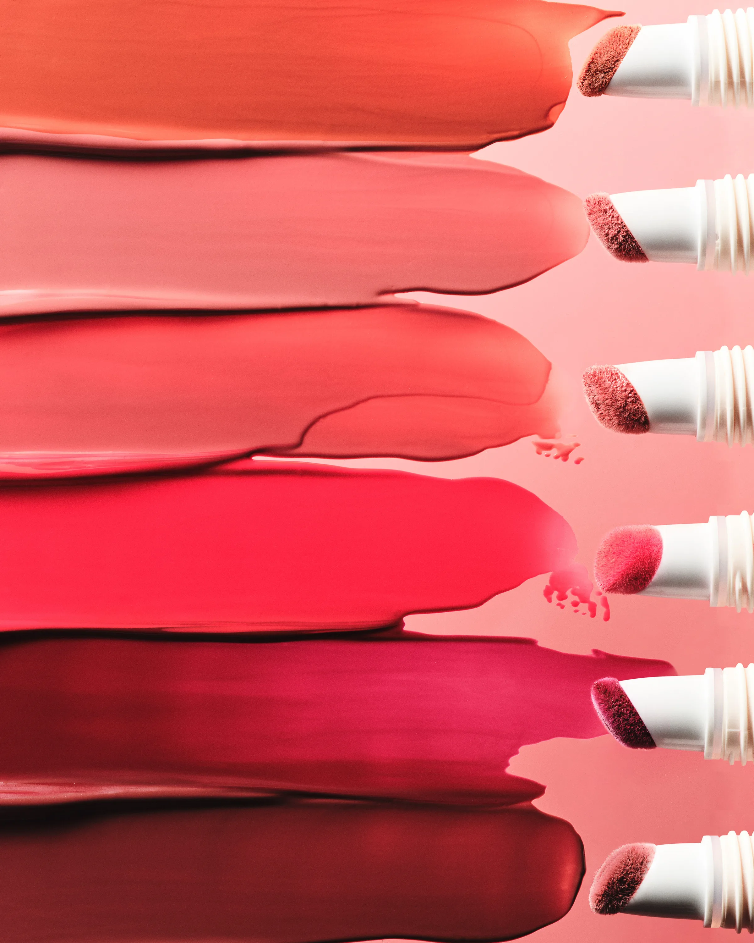 Six lip color swatches from coral to deep berry smeared beside white doe-foot applicators on pink, for Kylie Cosmetics.