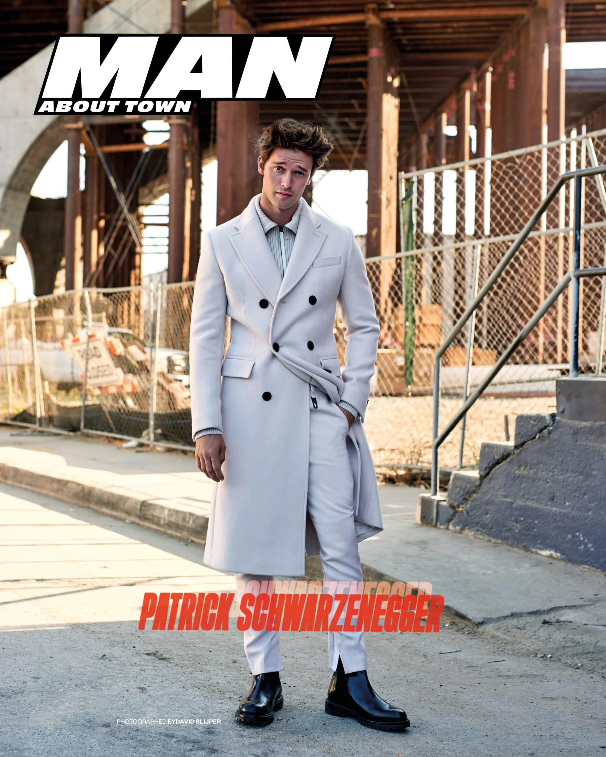 Magazine cover of Patrick Schwarzenegger in a white double-breasted coat under an elevated track, for Man About Town.