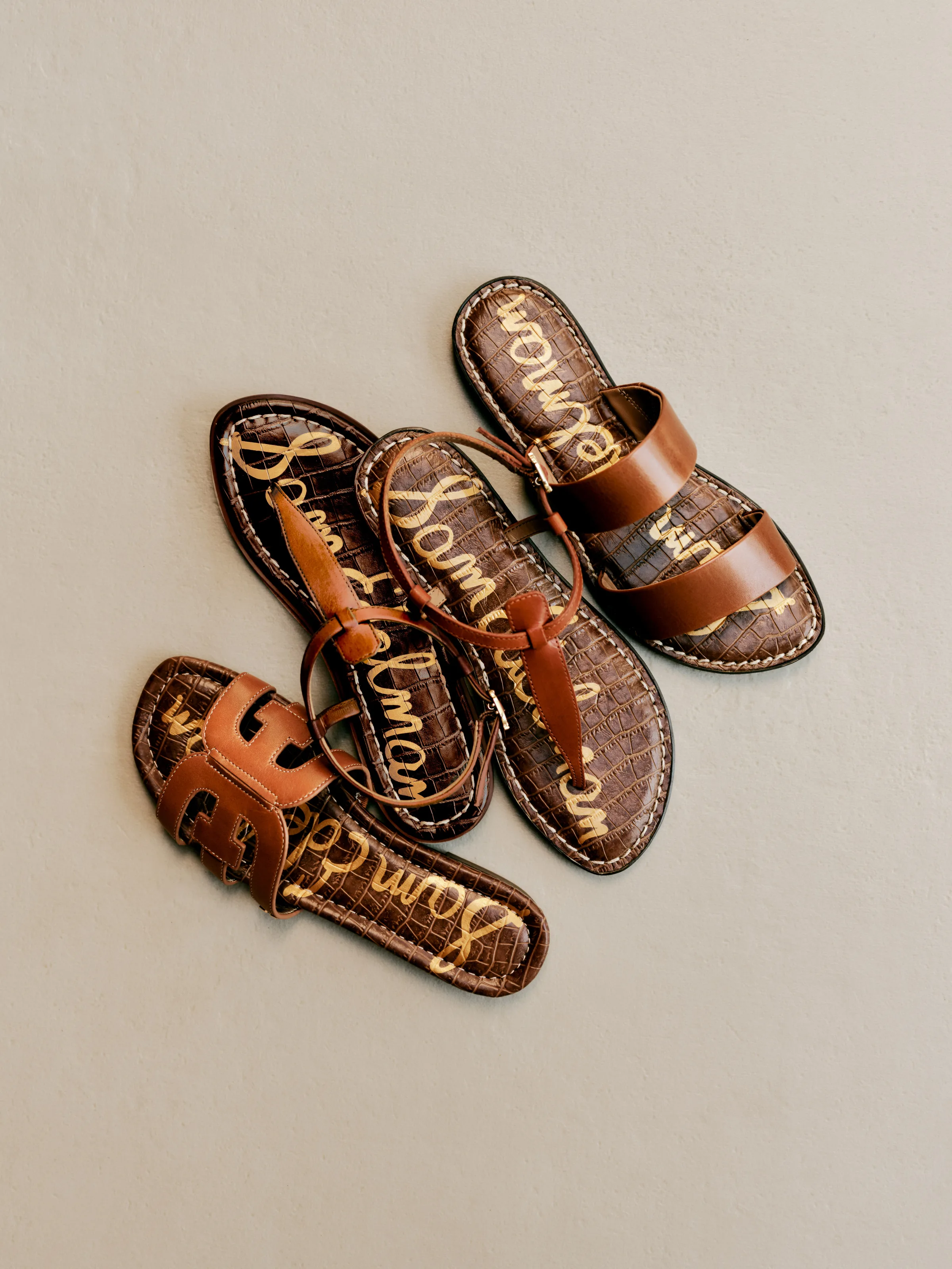 Three tan leather flat sandals with croc-embossed brown footbeds piled on a grey backdrop, for Sam Edelman.