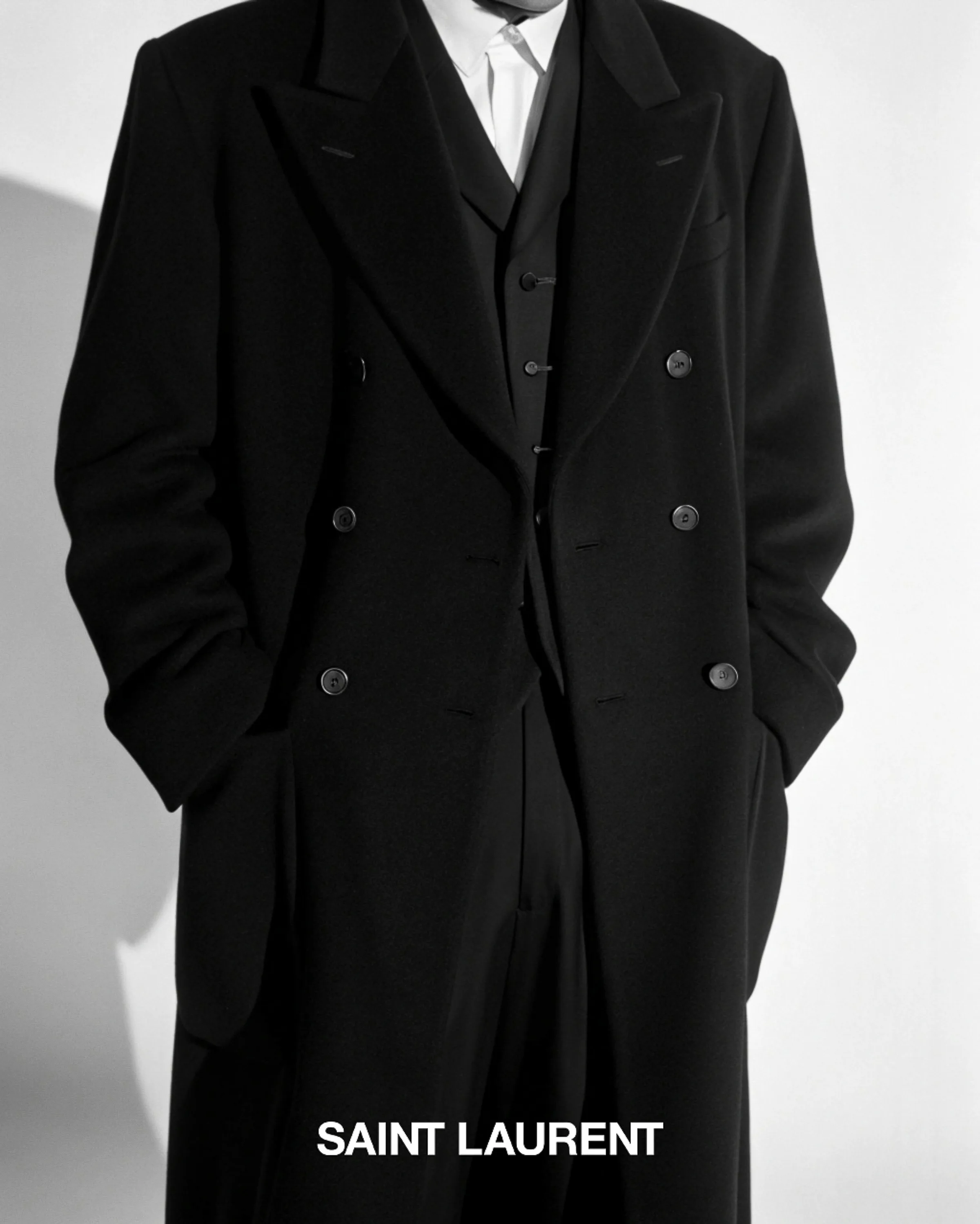 AI image of a model in a long black double-breasted overcoat and waistcoat against a white wall. Saint Laurent AI Concept.