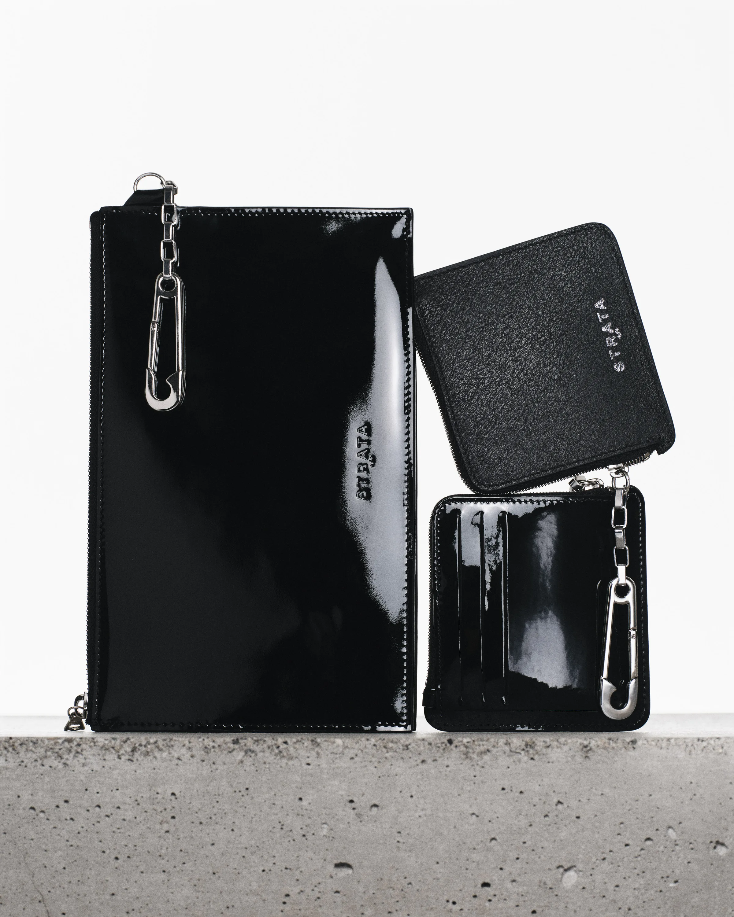Black patent and leather wallets with silver safety-pin clips propped on a concrete ledge, for Strata.