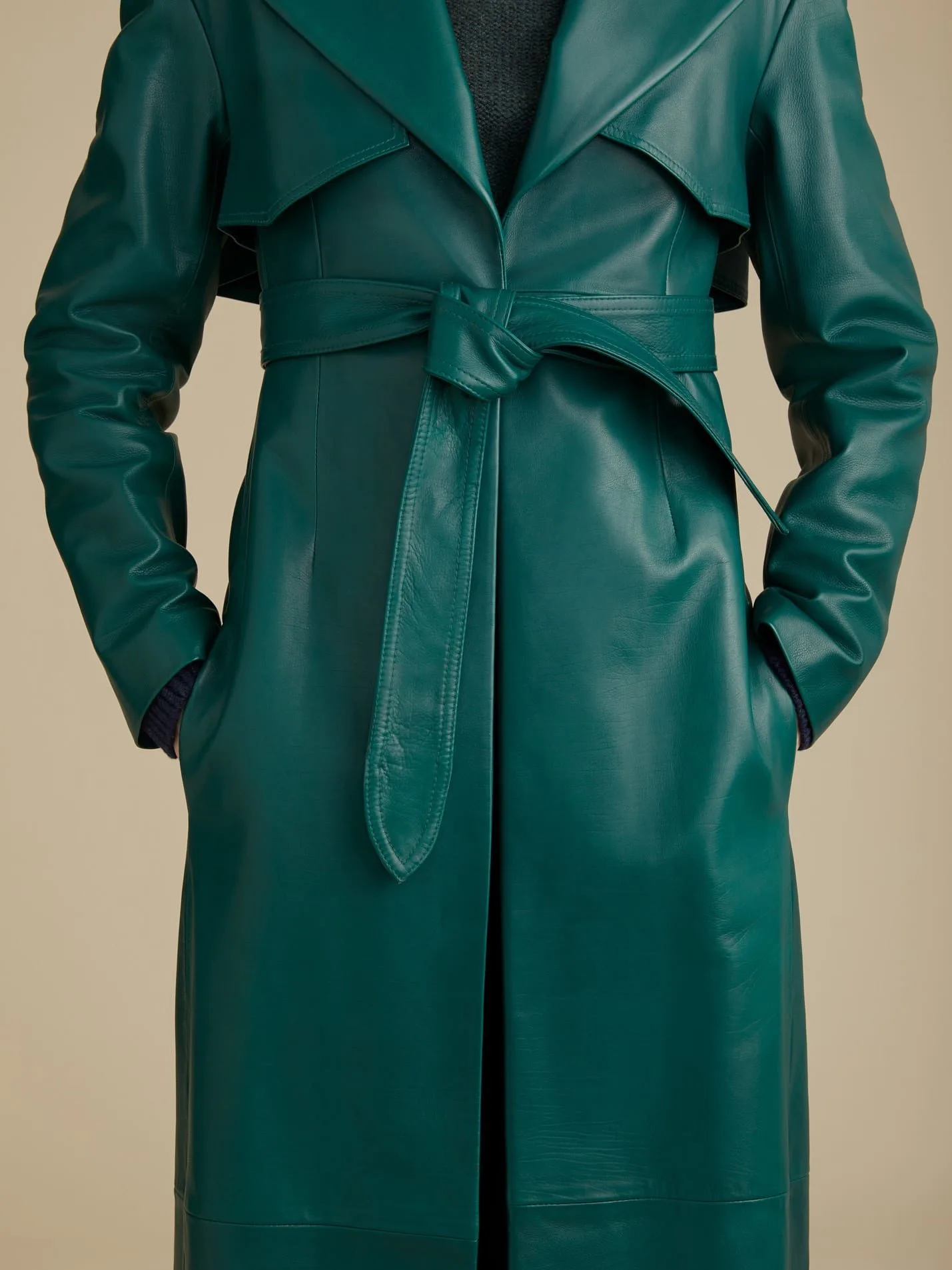 Model in a belted green leather trench coat with hands in pockets on a beige background, for Khaite.