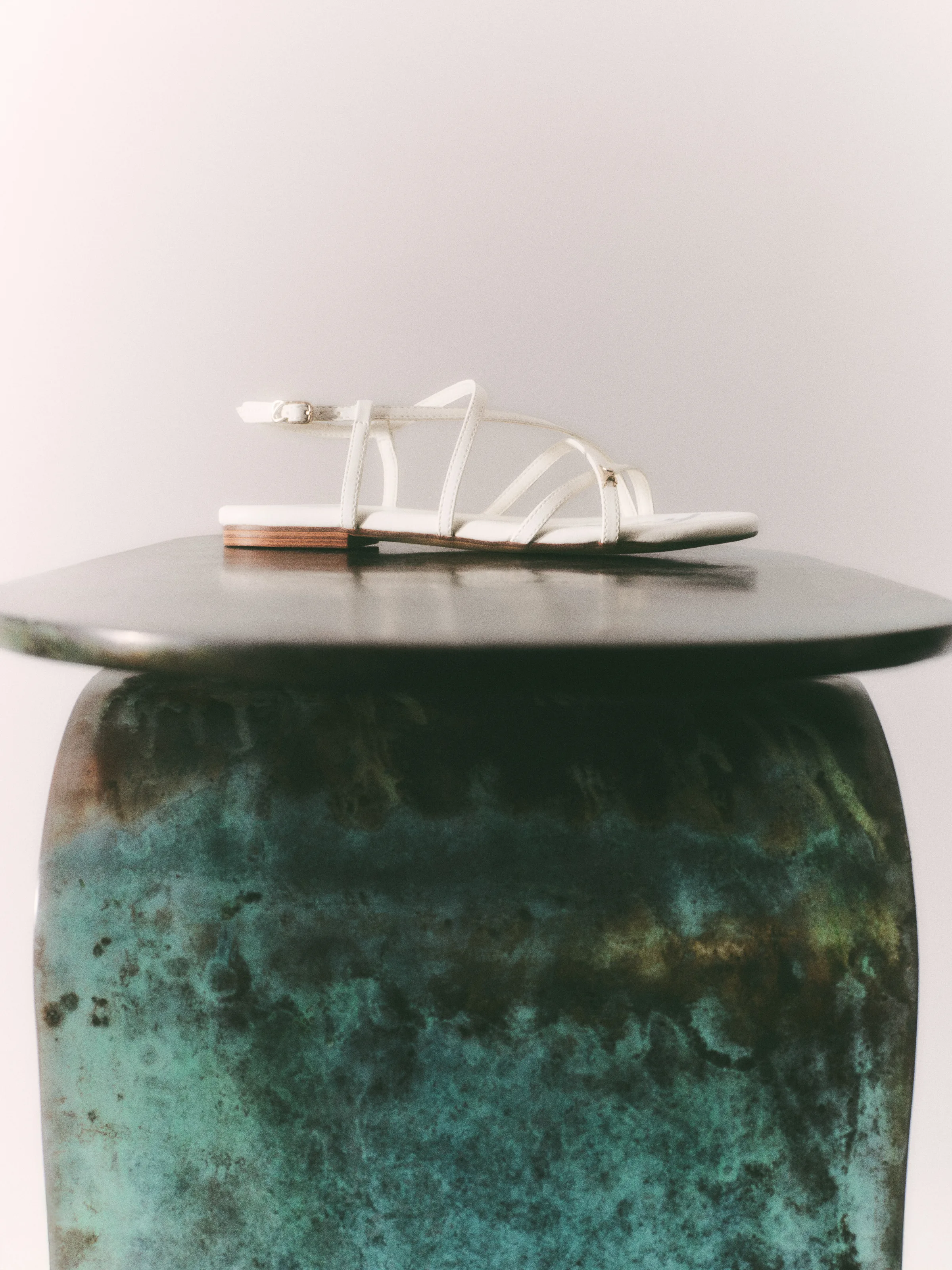 Bright white strappy flat sandal resting on a patinated green bronze pedestal, for Sam Edelman.