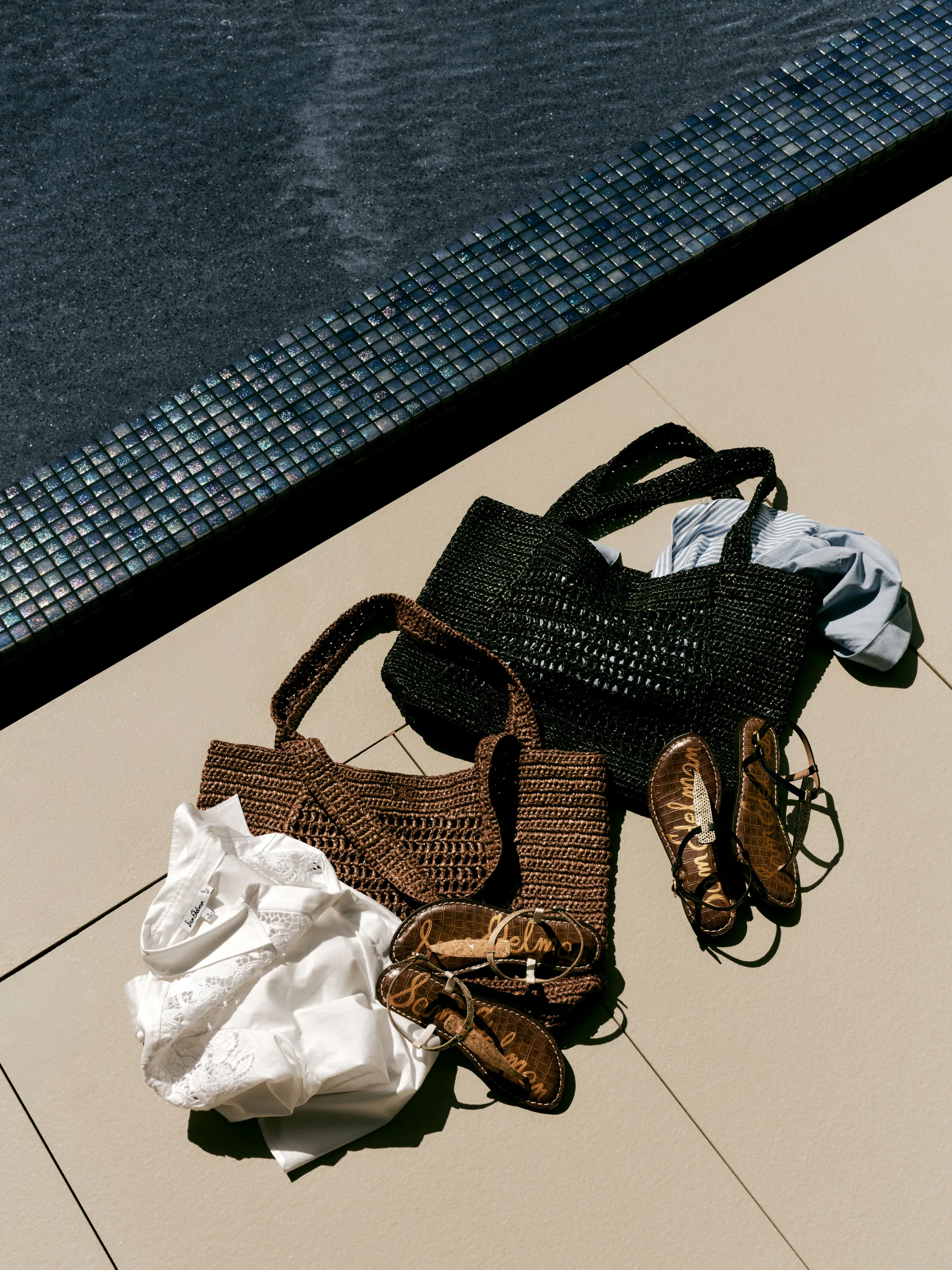 Black and brown woven raffia totes, white blouse and brown thong sandals beside a tiled pool edge, for Sam Edelman.