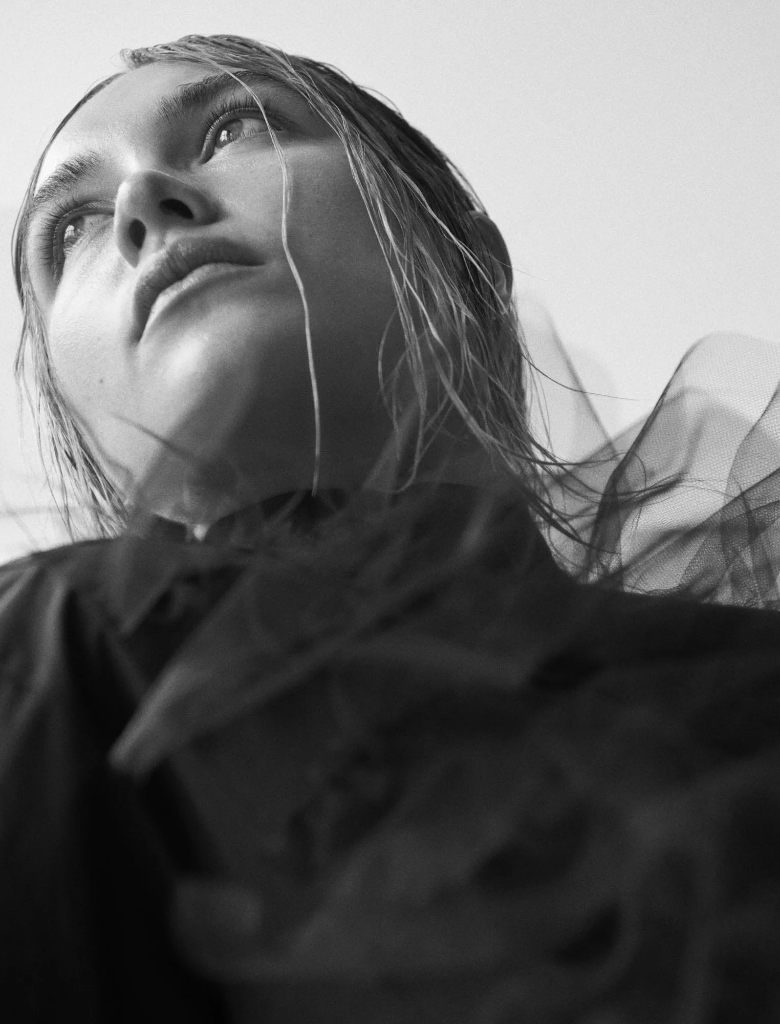 Black-and-white portrait of a model with wet slicked hair, dark shirt and sheer tulle, looking up, for The Last Magazine.