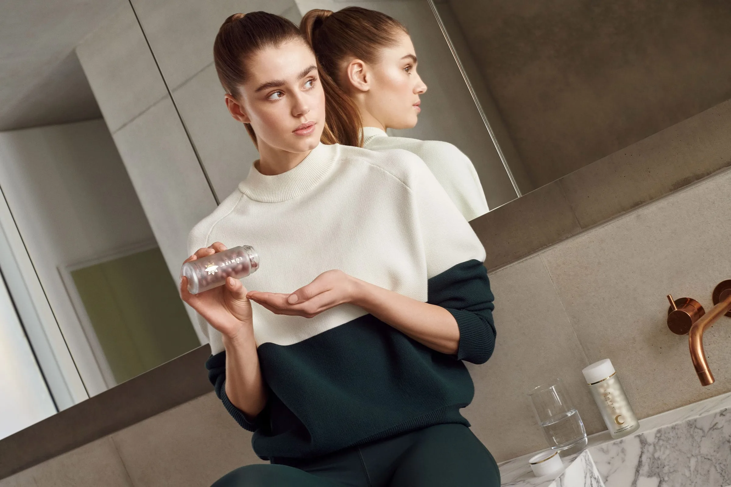 Model in a cream and green sweater holding a supplement bottle by a marble bathroom sink, for Net-a-Porter.