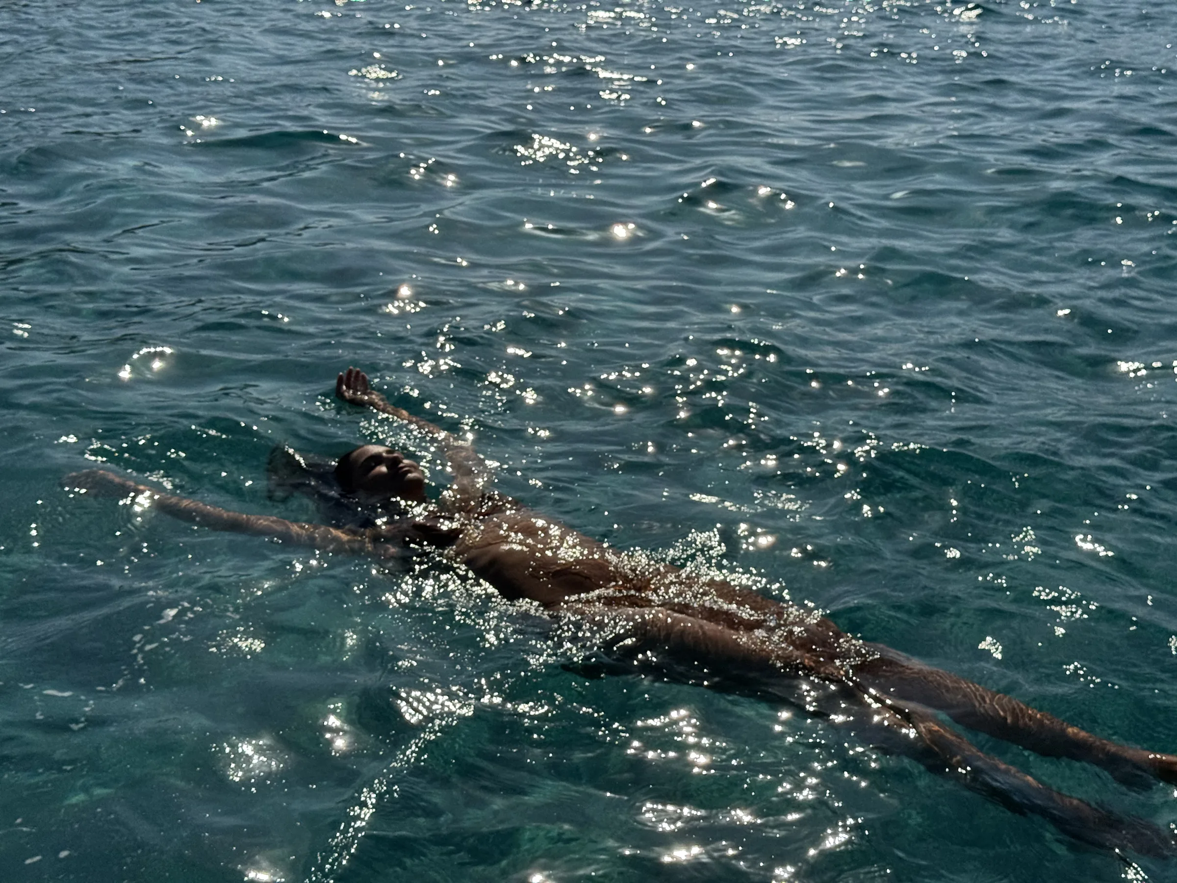 Woman floating on her back in sparkling turquoise sea water, arms outstretched in bright sunlight.