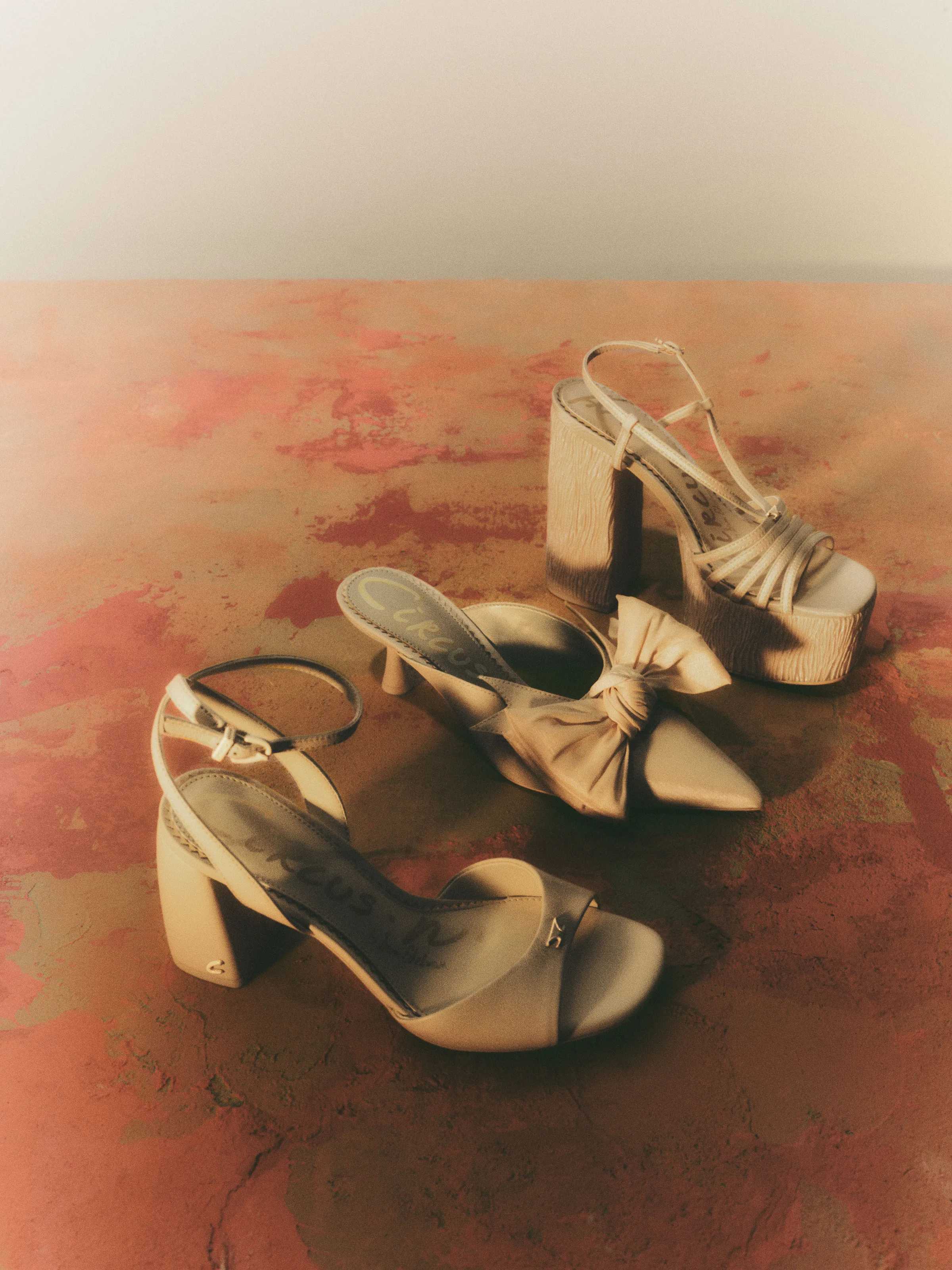 Three cream heels, a bow mule, ankle-strap block heel and platform sandal, on a mottled rust-red surface, for Circus NY.