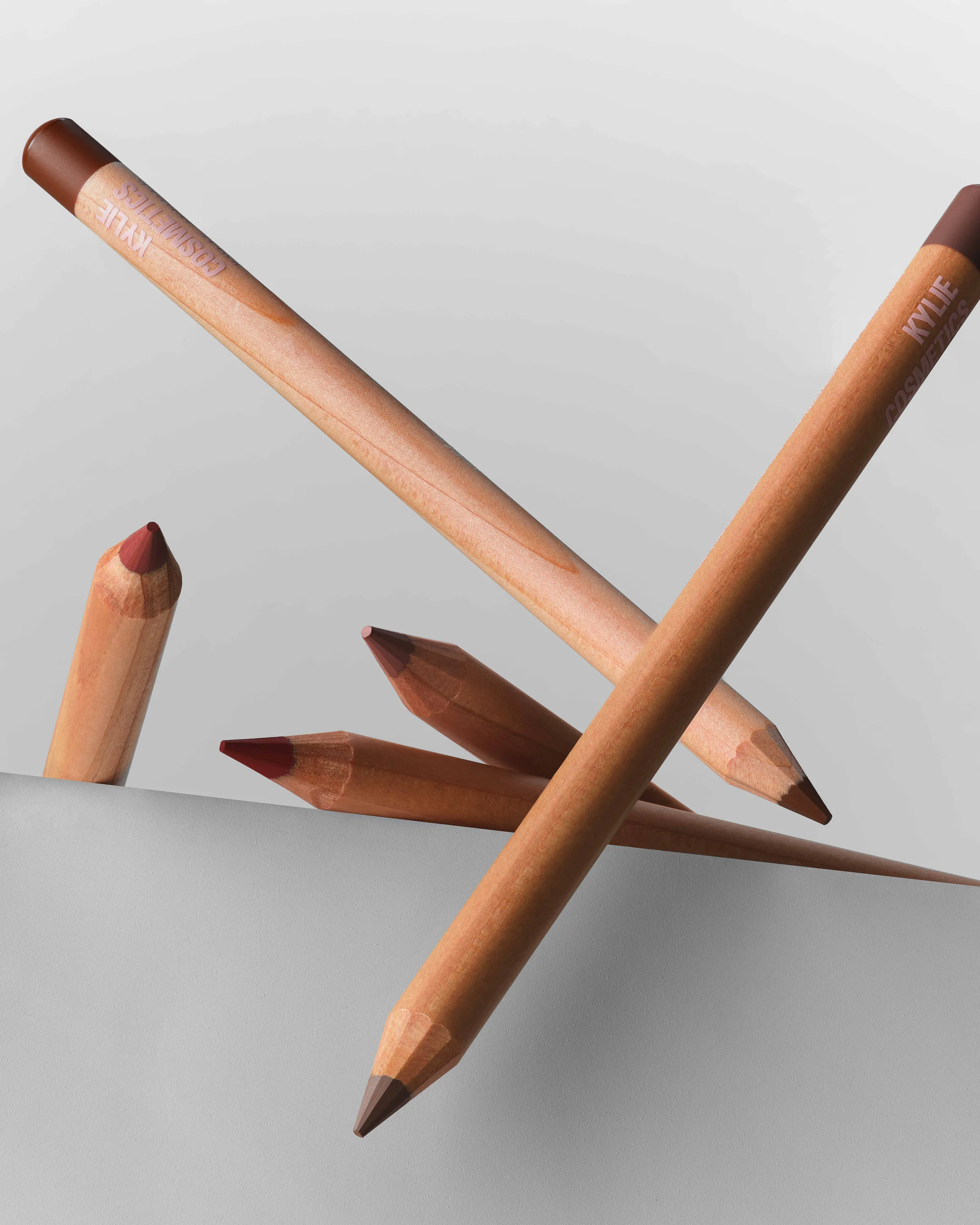 Wooden lip liner pencils in brown and red shades floating over a grey ledge, for Kylie Cosmetics.