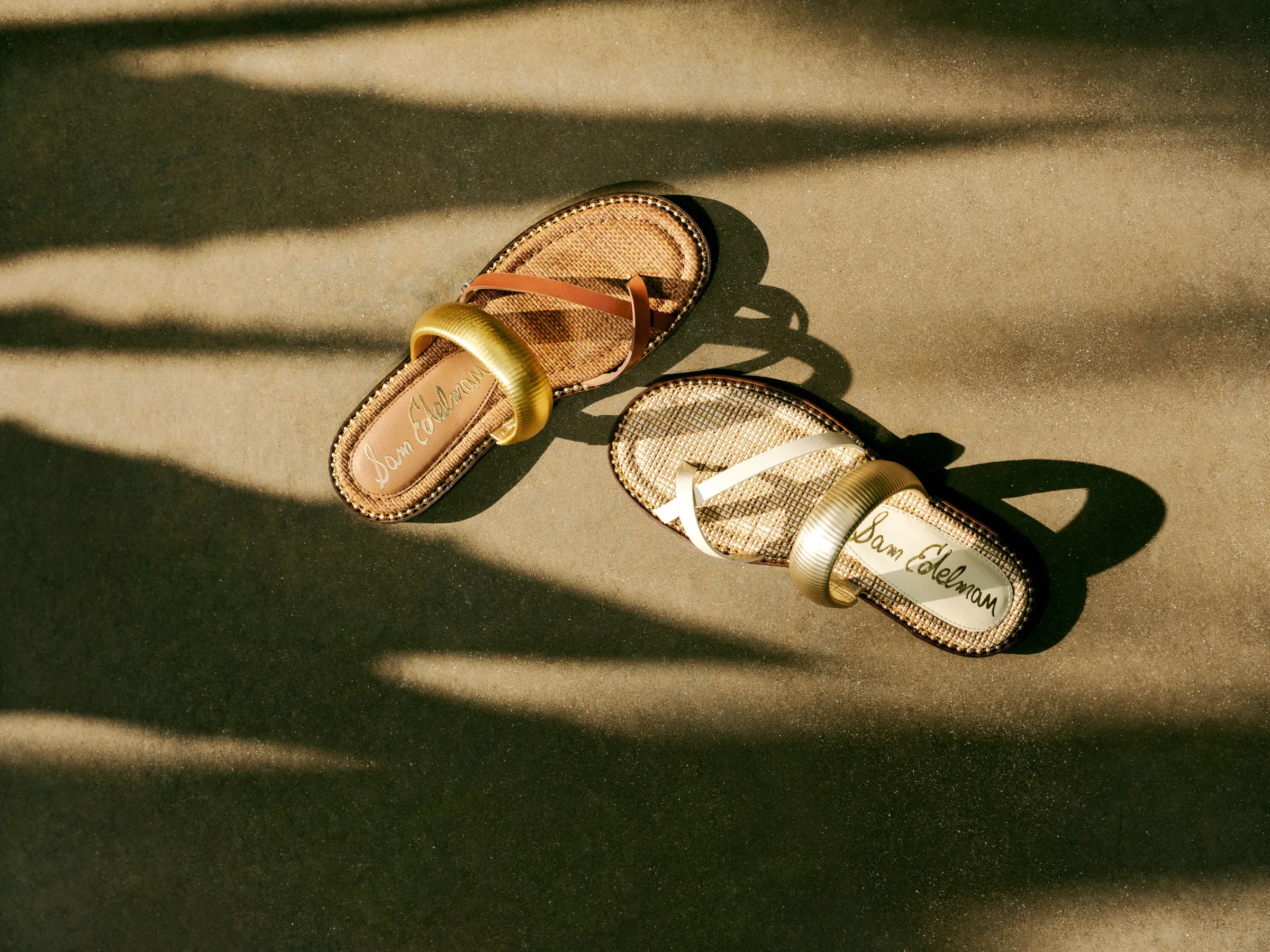 Two flat slide sandals with gold metallic straps and woven footbeds on a sunlit surface with palm shadows, for Sam Edelman.