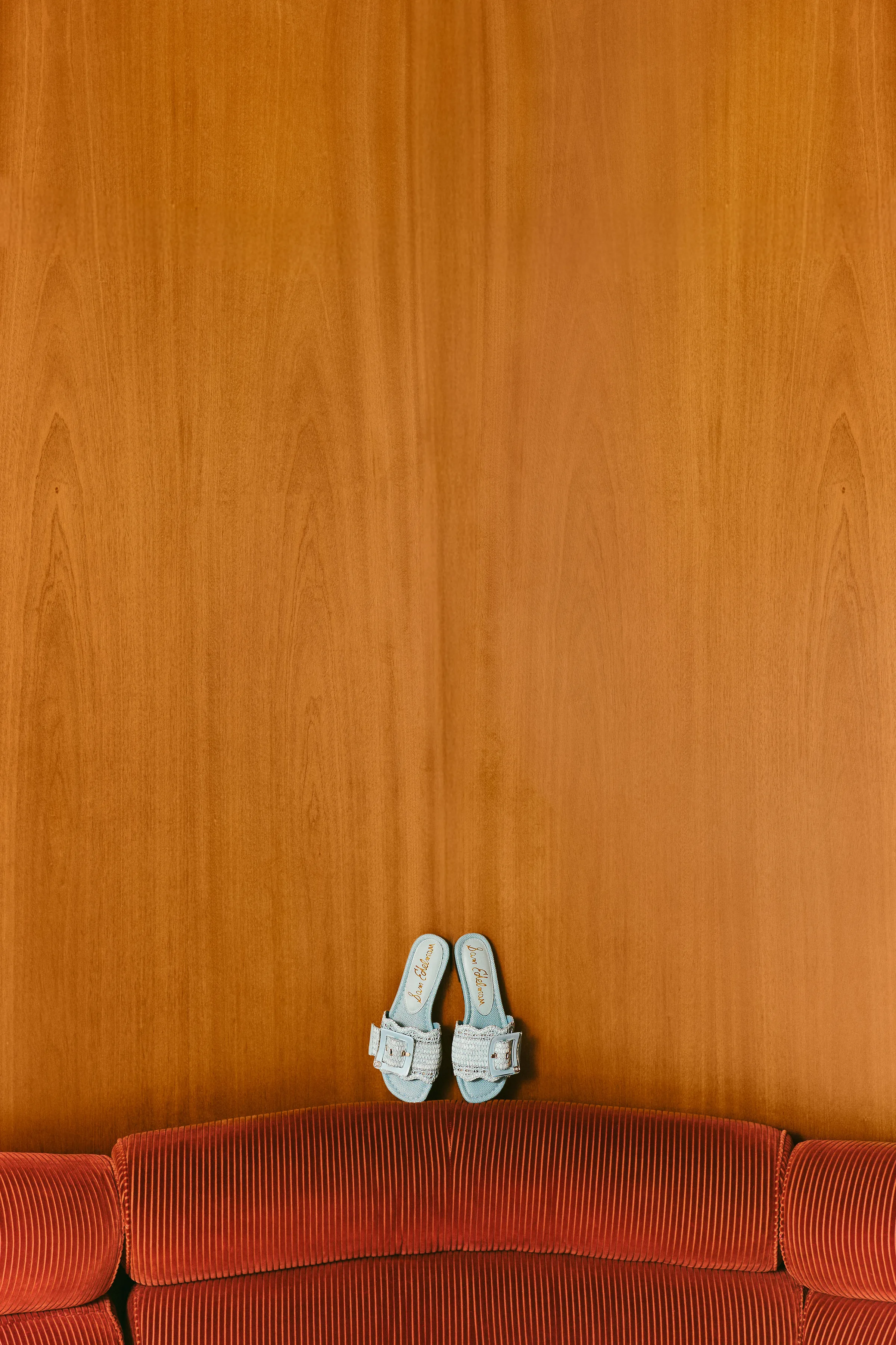 Light blue buckle slide sandals propped against a wood-panel wall above a rust corduroy sofa, for Sam Edelman.