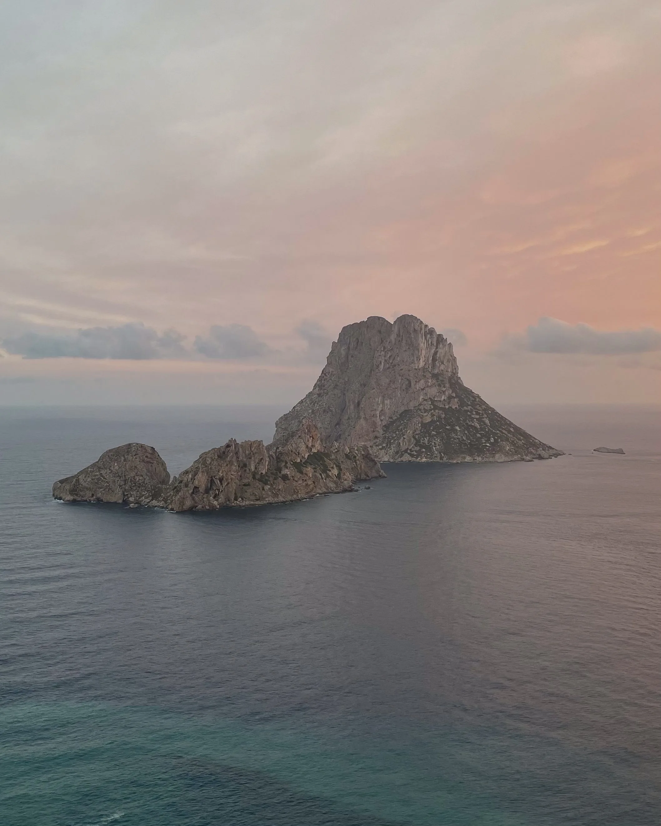 Rocky island rising from calm sea under a pink dusk sky, seen from a clifftop.
