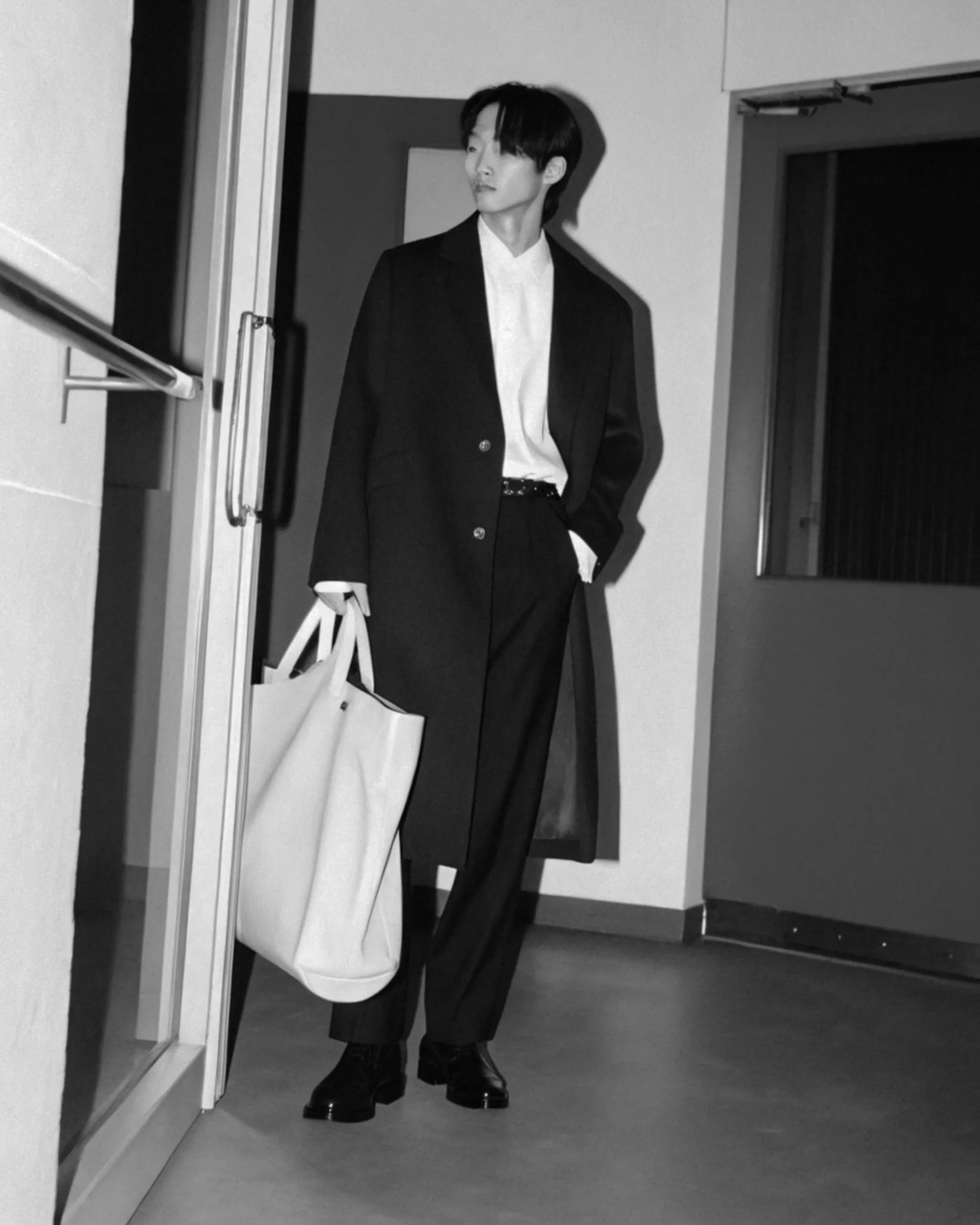 AI image of a model in a long black overcoat holding a white tote bag in a hallway, in black and white. Fursac AI Concept.